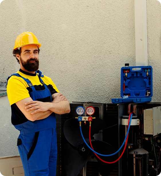 A male HVAC technician wearing a yellow hard hat and blue uniform with yellow accents, standing with arms crossed next to an HVAC unit with gauges and hoses.