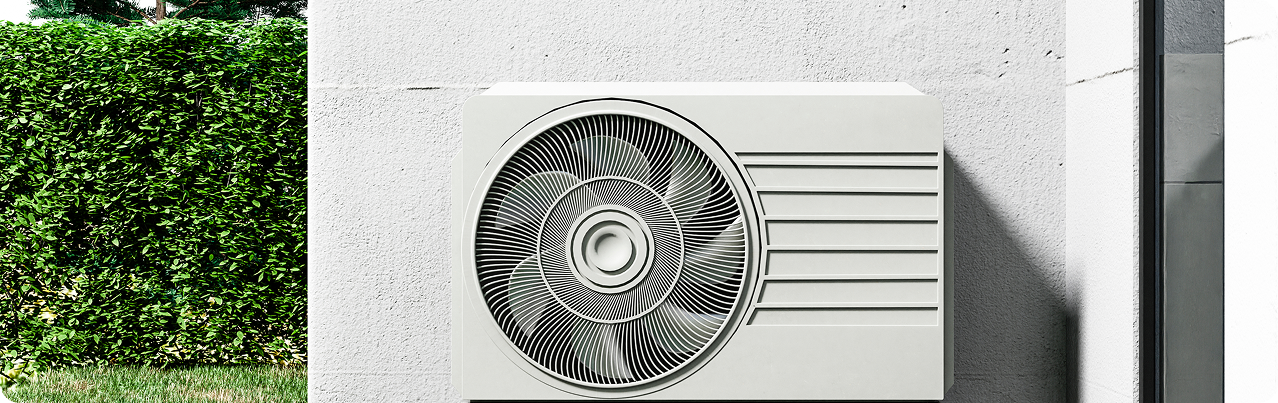 White outdoor air conditioning unit installed on a light gray wall beside a green hedge.