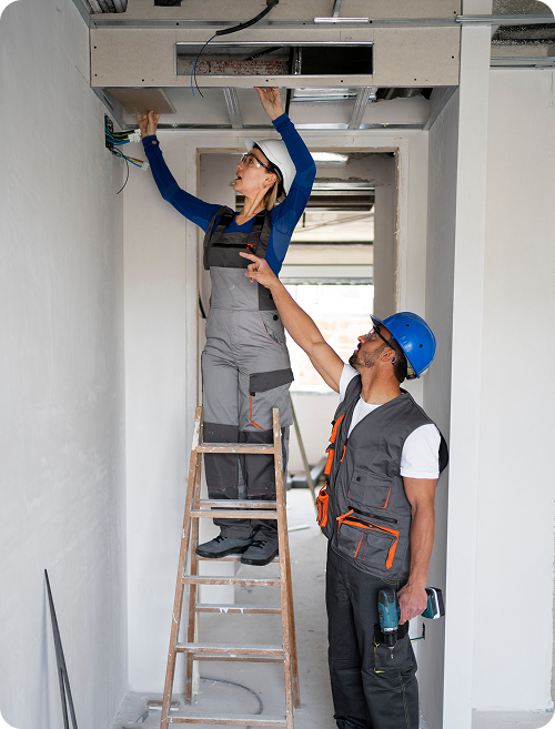 Two construction workers, a woman on a ladder and a man wearing a blue safety helmet, working together to install or repair ceiling wiring in a narrow hallway.