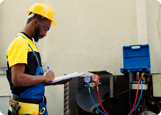 Electrician wearing a yellow safety helmet and yellow shirt, taking notes on a clipboard next to HVAC equipment with gauges and electrical connections.