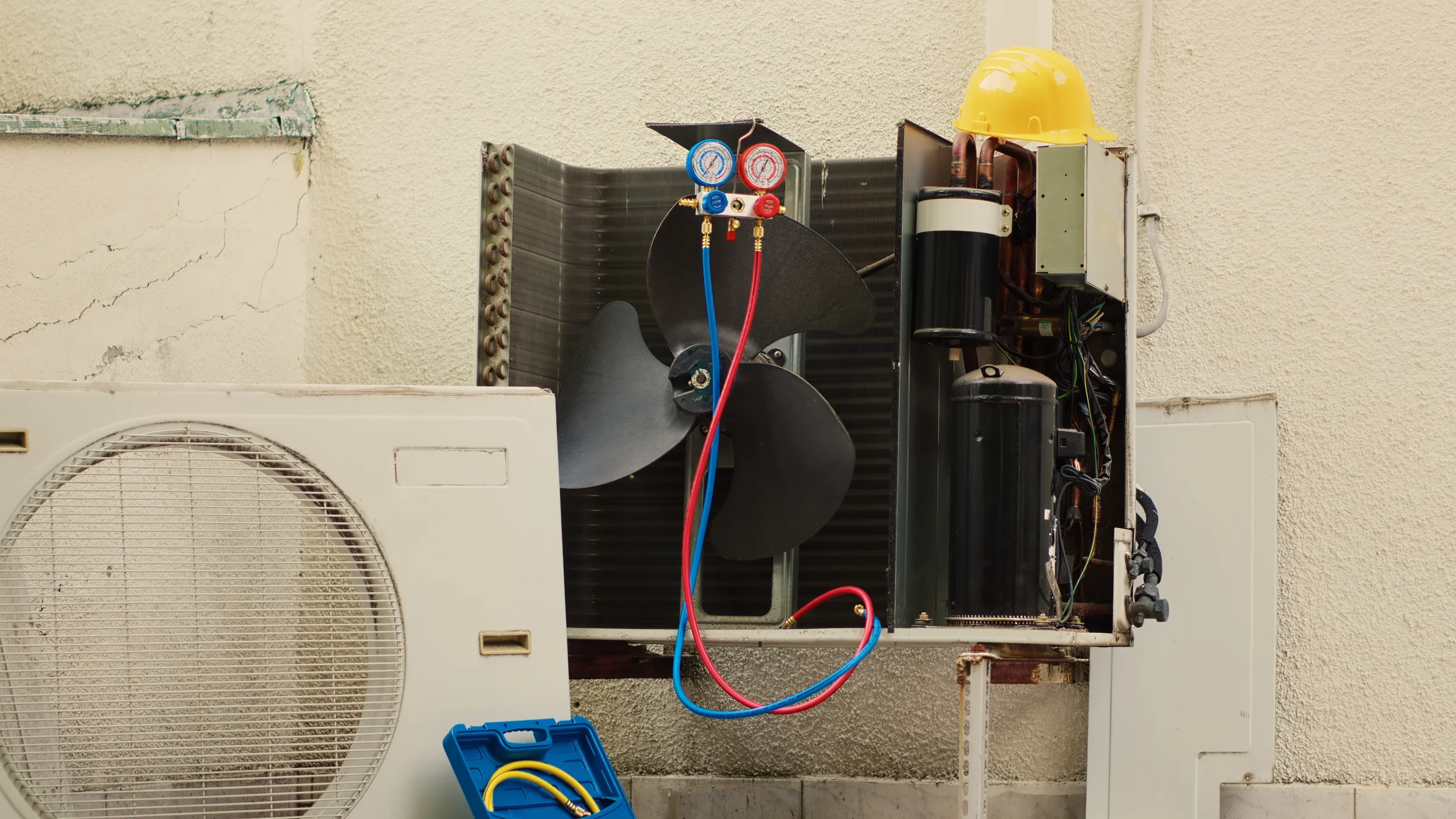Open outdoor air conditioning unit with exposed internal components, including a fan, compressor, and electrical wiring, mounted on a wall next to a white vent.