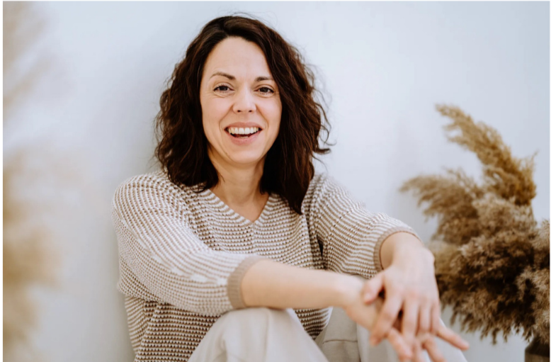 Lächelnde Frau mit braunen, lockigen Haaren in einem beige-gemusterten Pullover sitzt vor einer weißen Wand, umgeben von Trockenblumen.