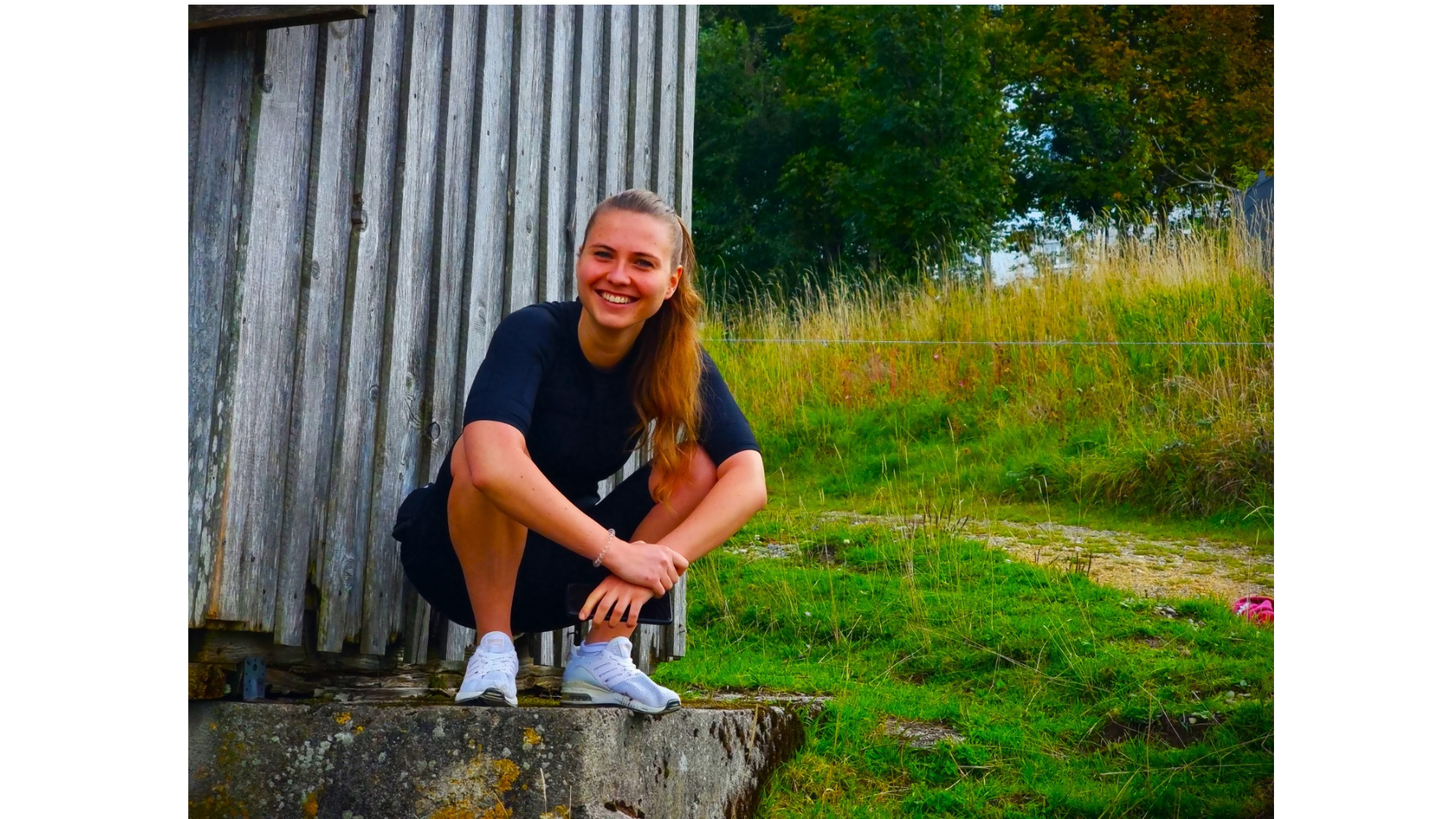 Junge Frau mit roten Haaren in schwarzem T-Shirt und Turnschuhen lächelnd, sitzend auf einem alten Baumstumpf neben einem holzverschalten Gebäude in einem Grünen Außenbereich