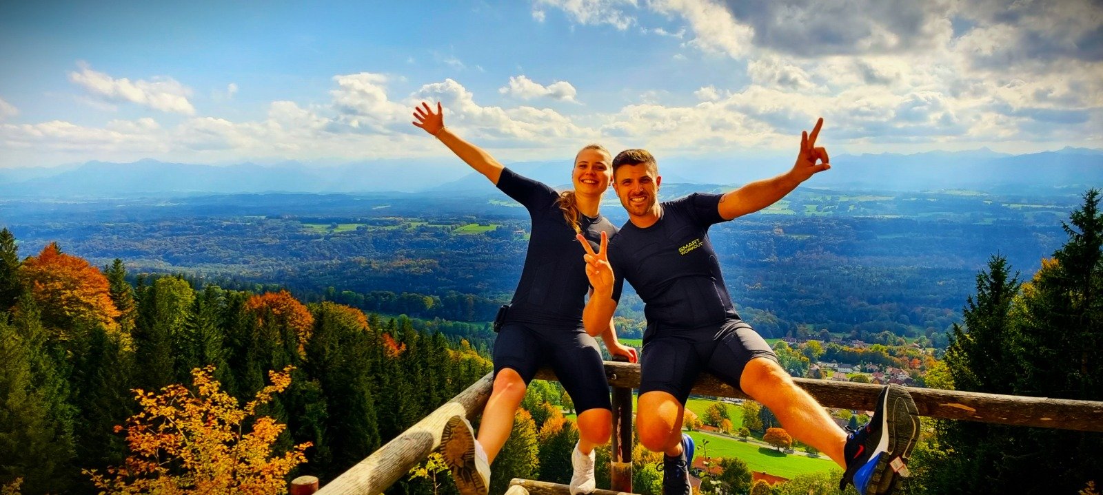 Ein lachendes Paar in sportlicher Kleidung sitzt auf einem Holzgeländer auf einem Berg mit Blick auf eine Herbstlandschaft, blauen Himmel und Wolken im Hintergrund, macht eine Peace-Zeichen-Geste und genießt die Aussicht.