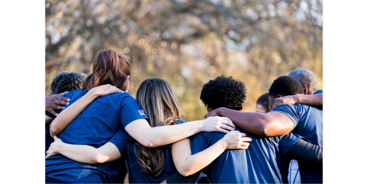 Eine Gruppe von Menschen in blauen T-Shirts steht im Kreis und umarmt sich bei Sonnenuntergang im Freien.