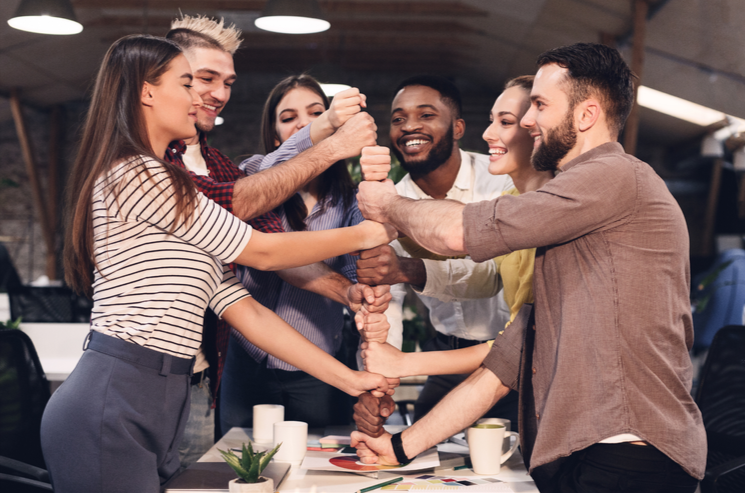 Sechs Menschen in einem Büro, die sich freundschaftlich an den Händen fassen und sich die Hände zu einem gegenseitigen Fauststoß klopfen, lachen und sich freuen.