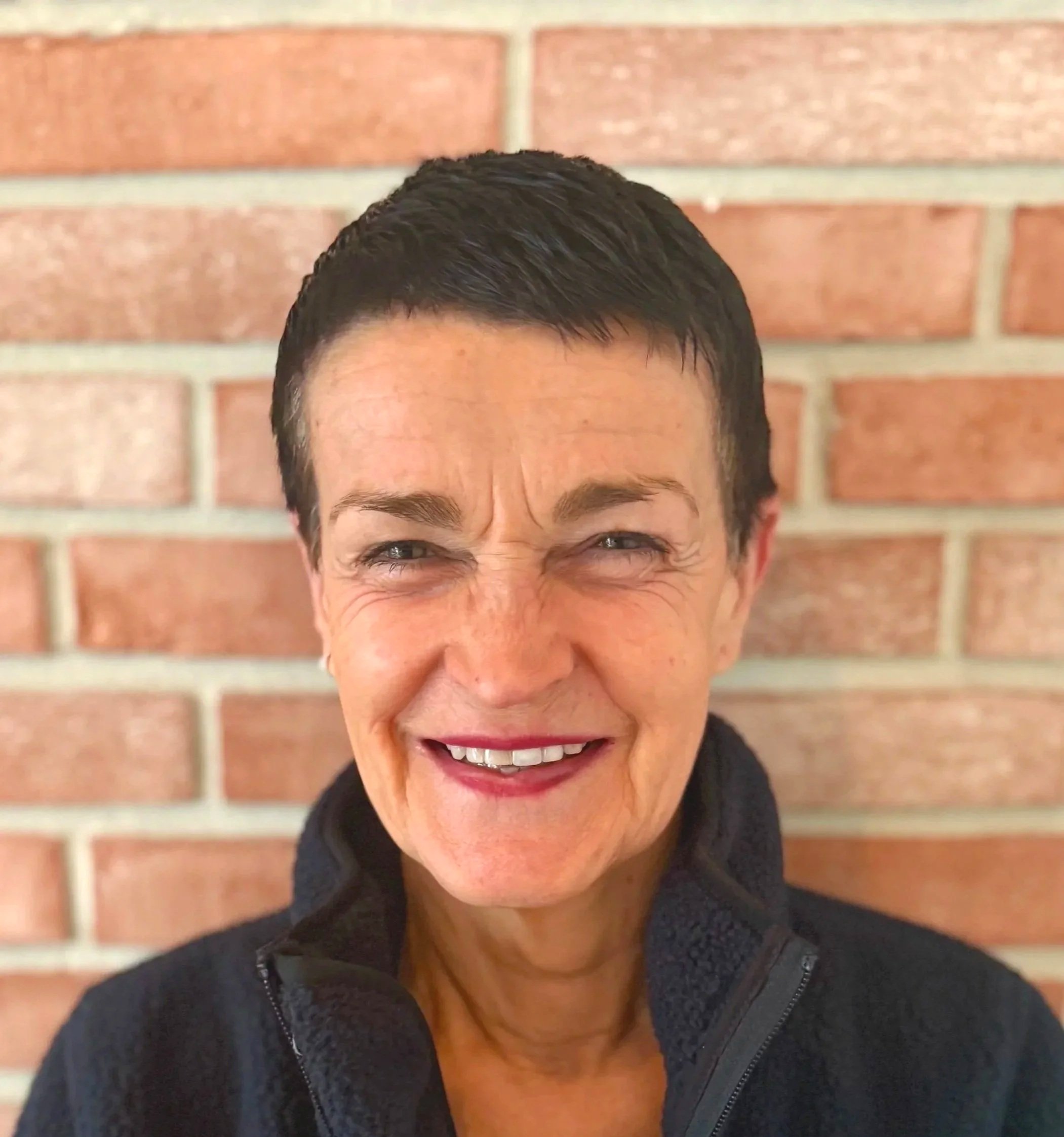 Close-up of a smiling woman with short dark hair, wearing a dark jacket, standing in front of a brick wall.