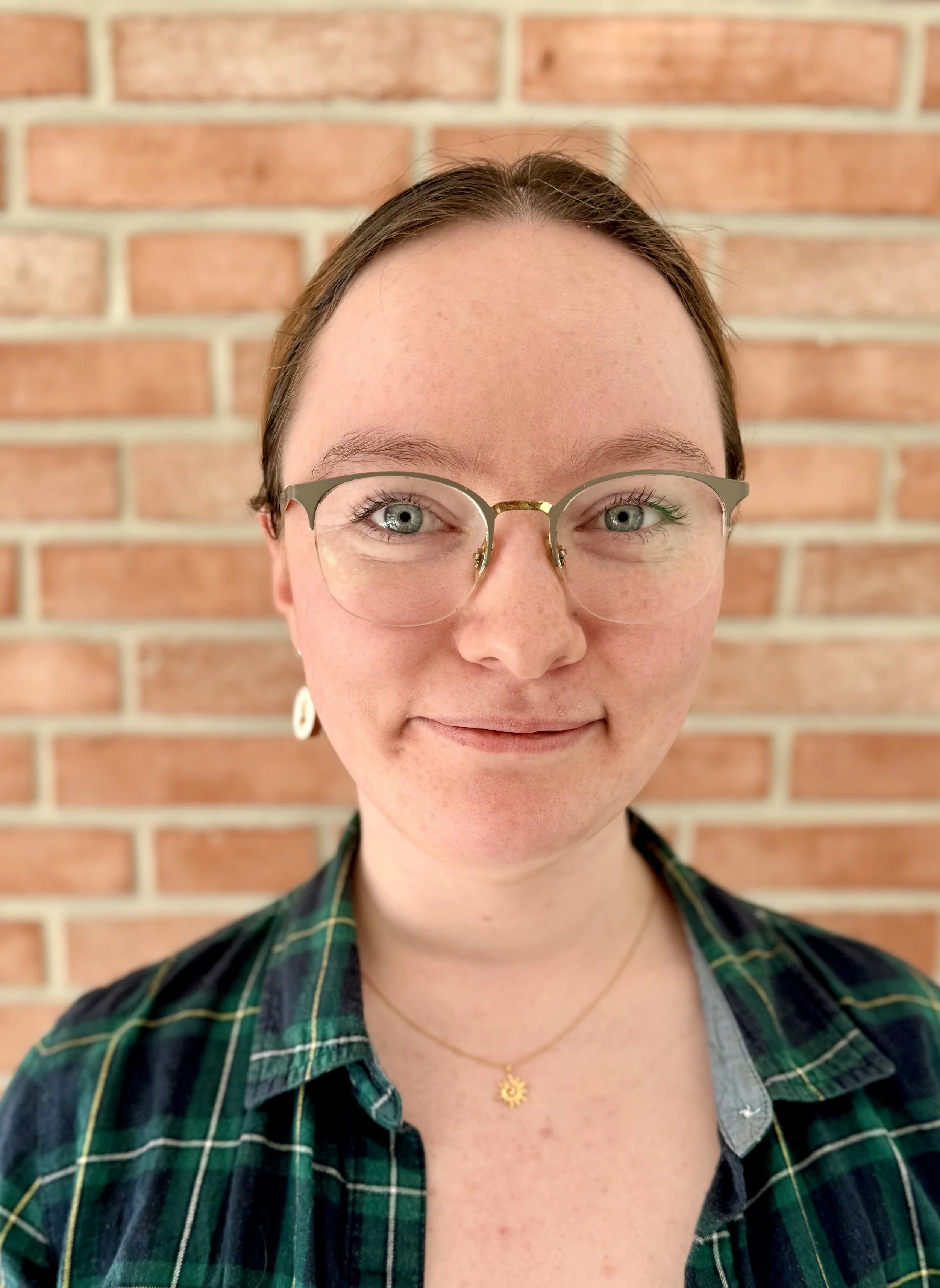 Close-up of a smiling woman with blue eyes, glasses, and earrings in front of a brick wall.