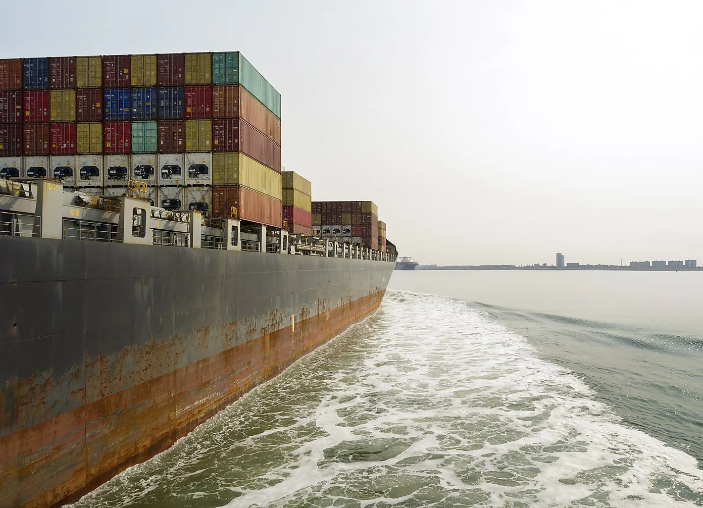 Cargo ship with stacked shipping containers sailing in calm water near a distant city skyline.
