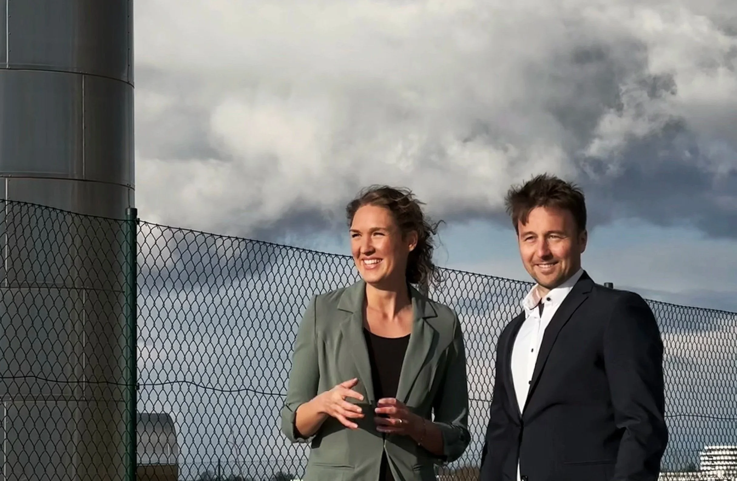 Ein Mann und eine Frau stehen im Freien vor einem Zaun, beide sind am Lächeln. Es ist bewölkt am Himmel.