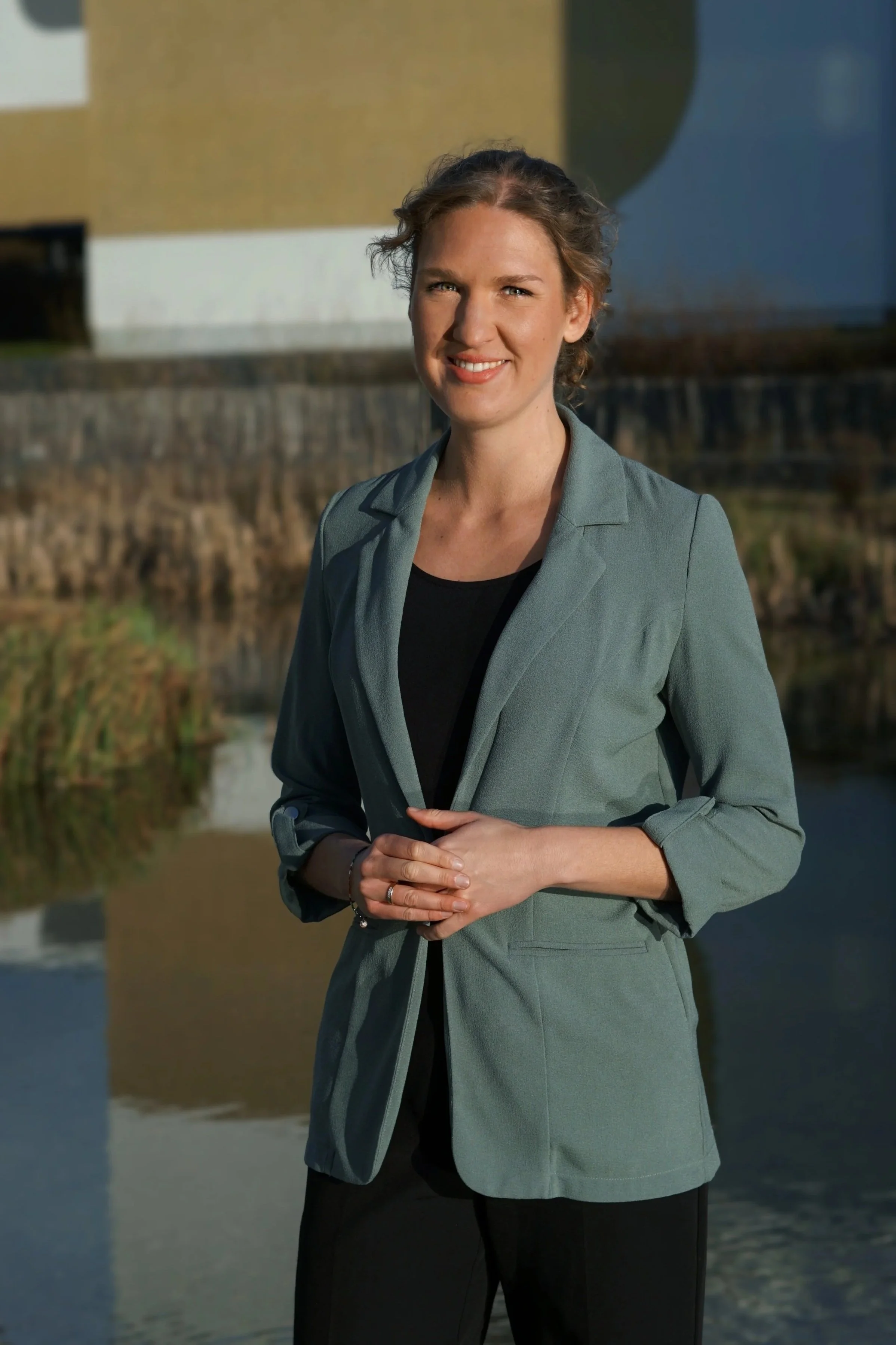Eine Frau trägt eine graue Blazer vor einem Wasserkanal mit Gebäuden im Hintergrund.