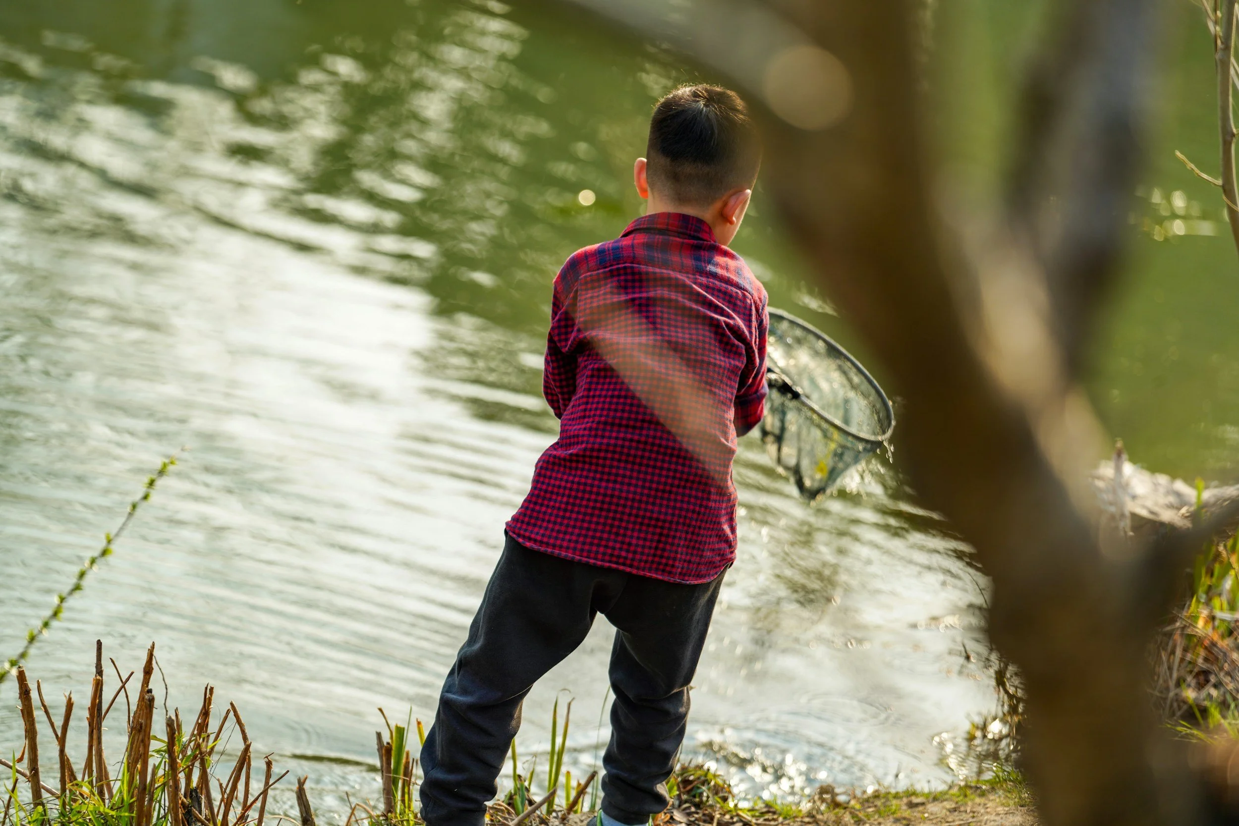 A young boy with short dark hair, wearing a red plaid shirt and dark pants, fishing in a shallow river surrounded by green trees.