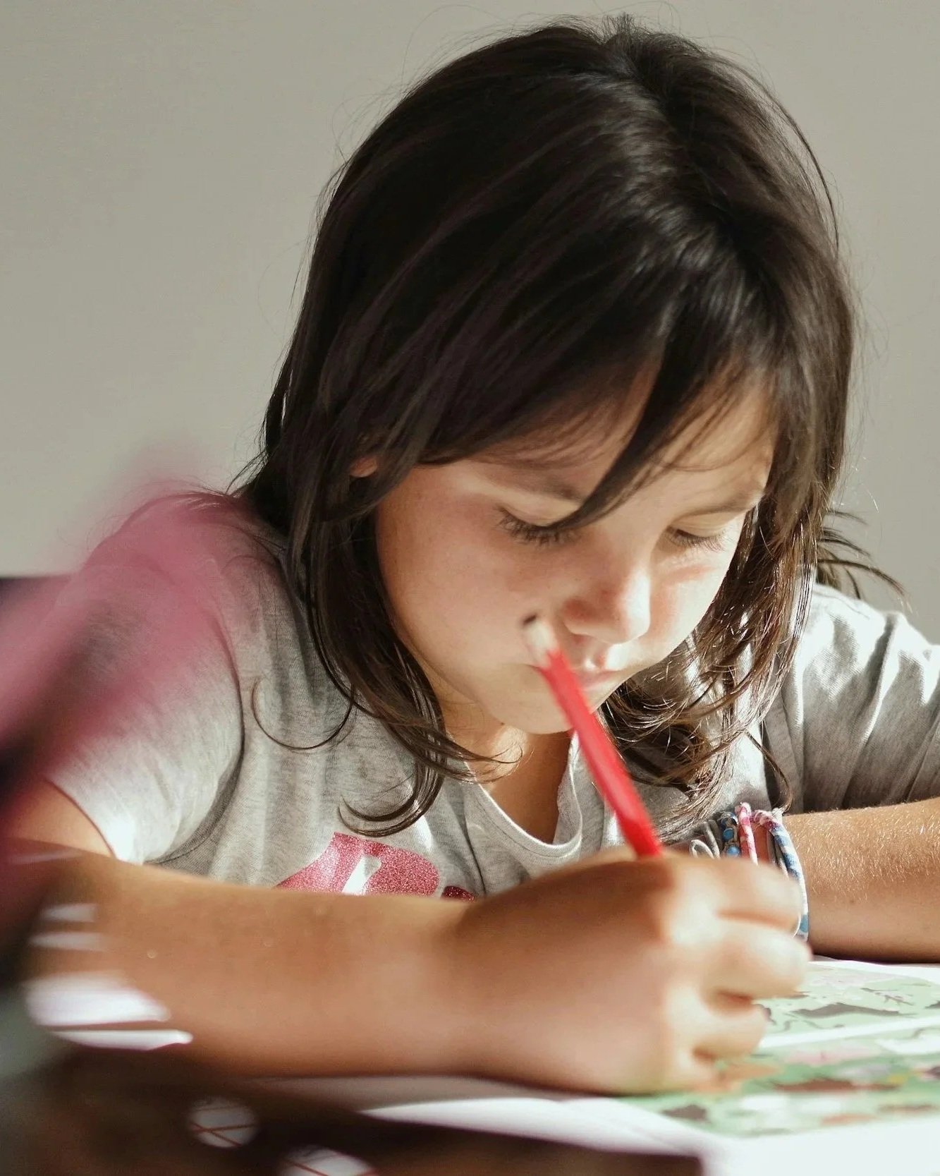 A young girl with brown hair is coloring or drawing with a red crayon or marker at a table.