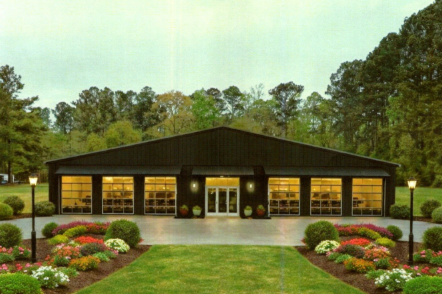 A single-story modern commercial building with several large garage-style windows and a central entrance, surrounded by a well-maintained garden with flower beds and green grass, with tall trees in the background.