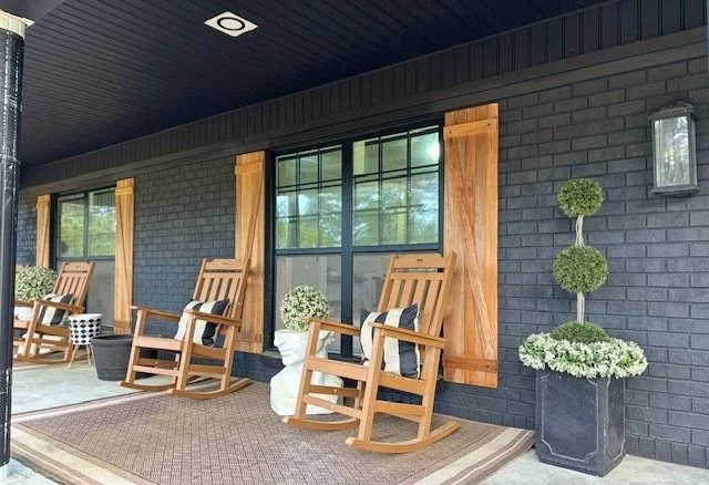 Porch with wooden rocking chairs, potted plants, window with wooden shutters, and wall-mounted light fixture.