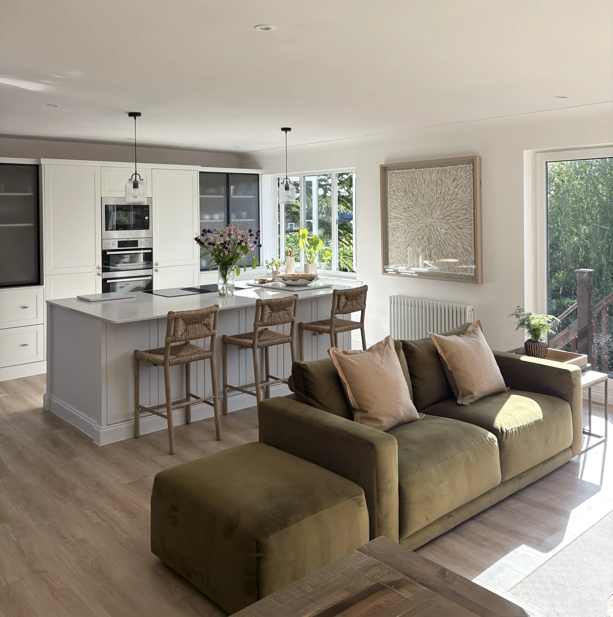 Modern kitchen with white cabinets, stainless steel oven, and large windows with views of greenery, featuring a kitchen island with a vase of pink flowers and brown woven bar stools.