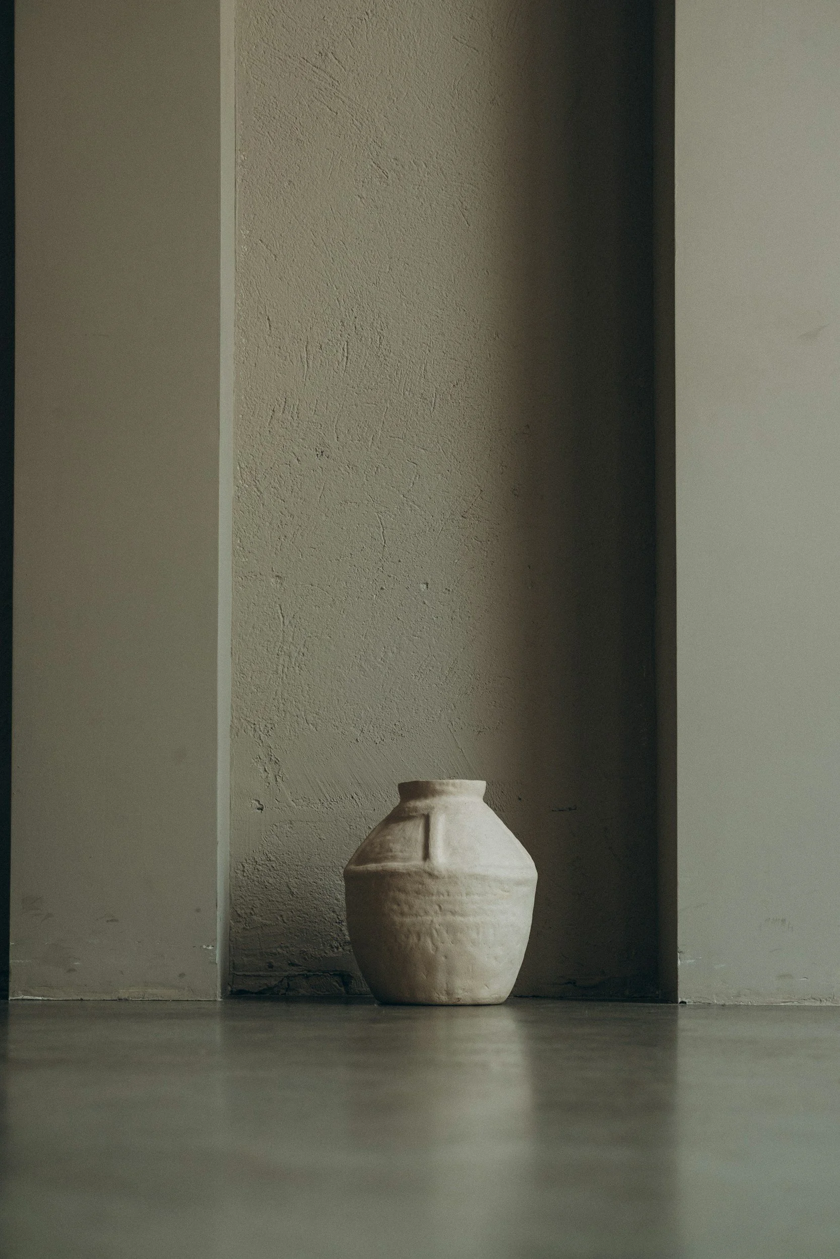 A large, light-colored ceramic vase with a rounded body and narrow opening, placed on a dark floor against a textured wall in a minimalistic setting.