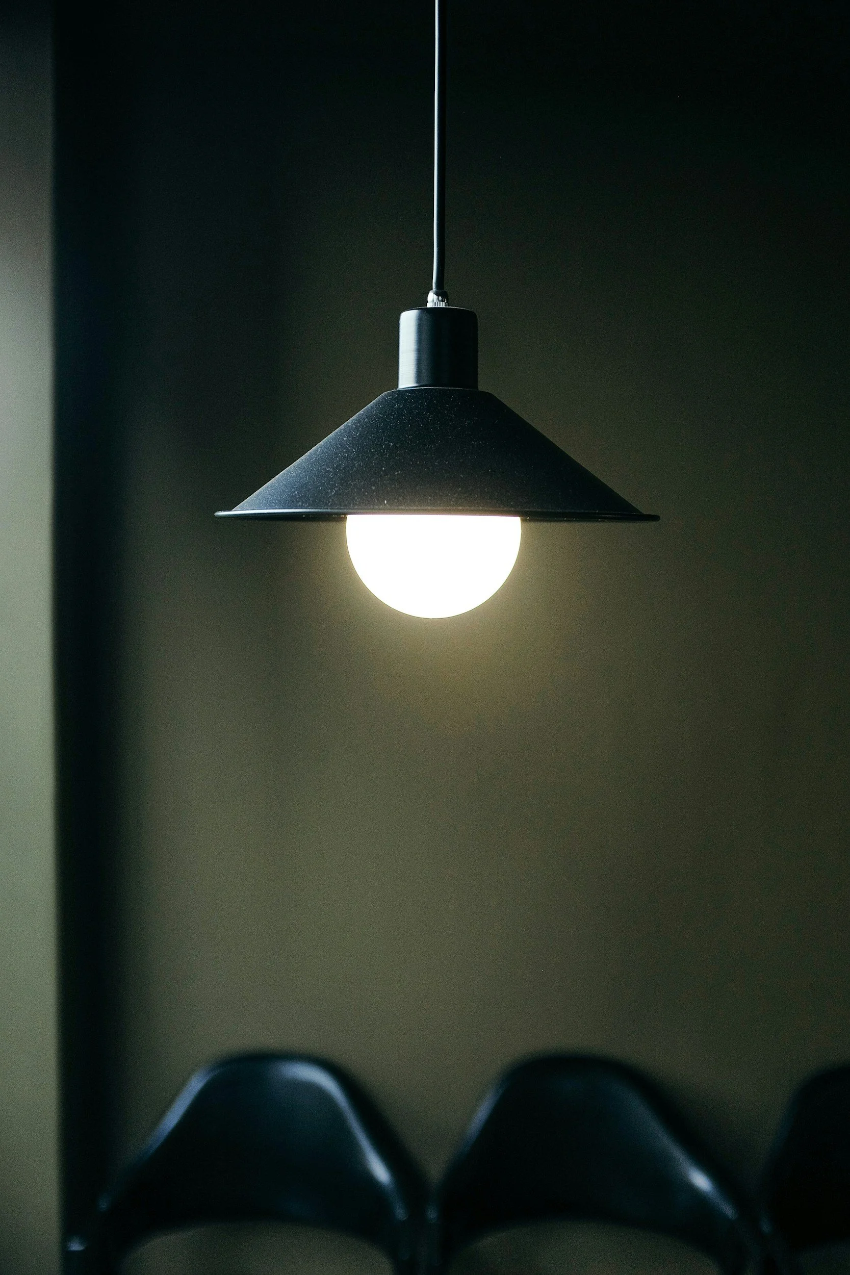 A black hanging pendant light with a round light bulb, viewed from below, against a dark wall, with a pair of shoes visible at the bottom of the image.