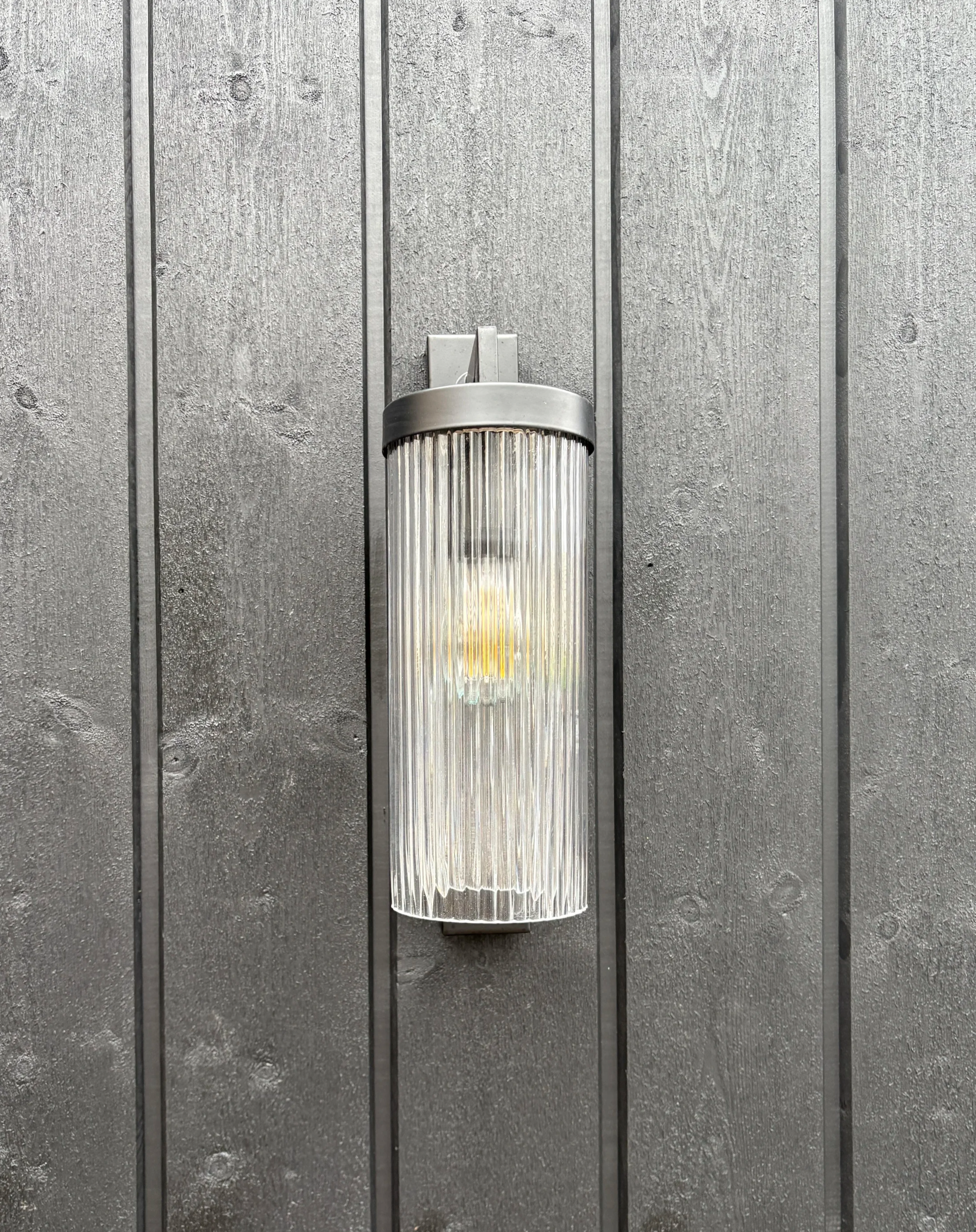 Wall-mounted light fixture with cylindrical, ribbed glass cover and a black metal top, mounted on a black timber architectural cladding.