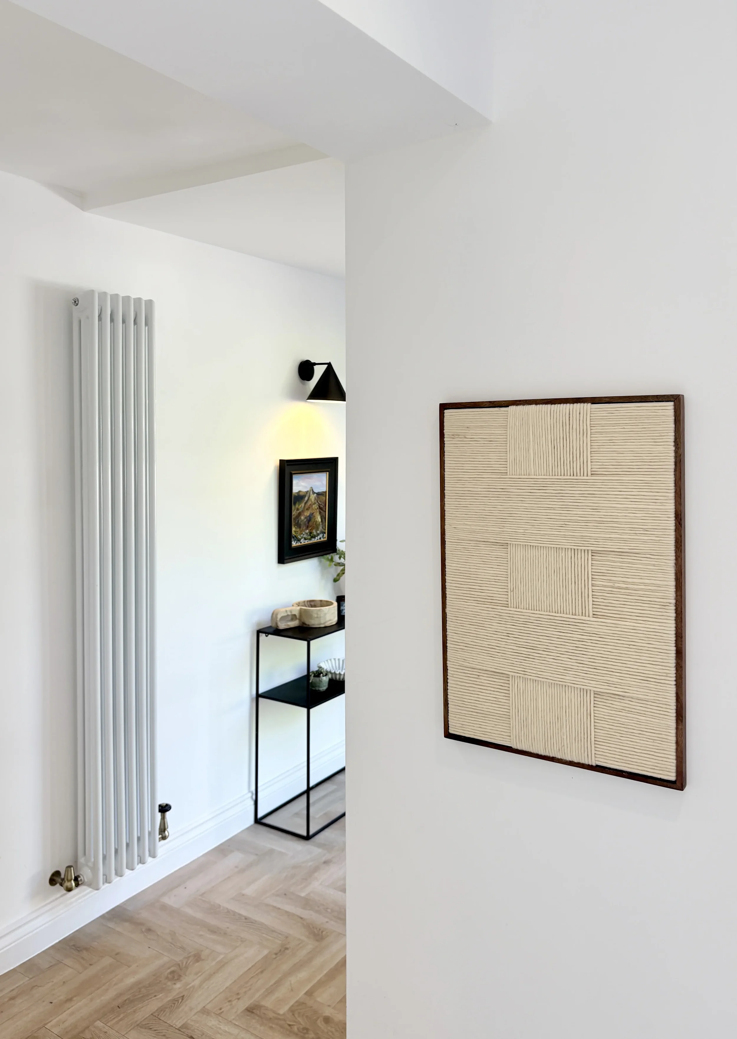 Interior view of a modern, minimalist living space with a white wall, beige textured wall art, narrow black metal console table with decorative items, a built-in white radiator, and hardwood flooring.