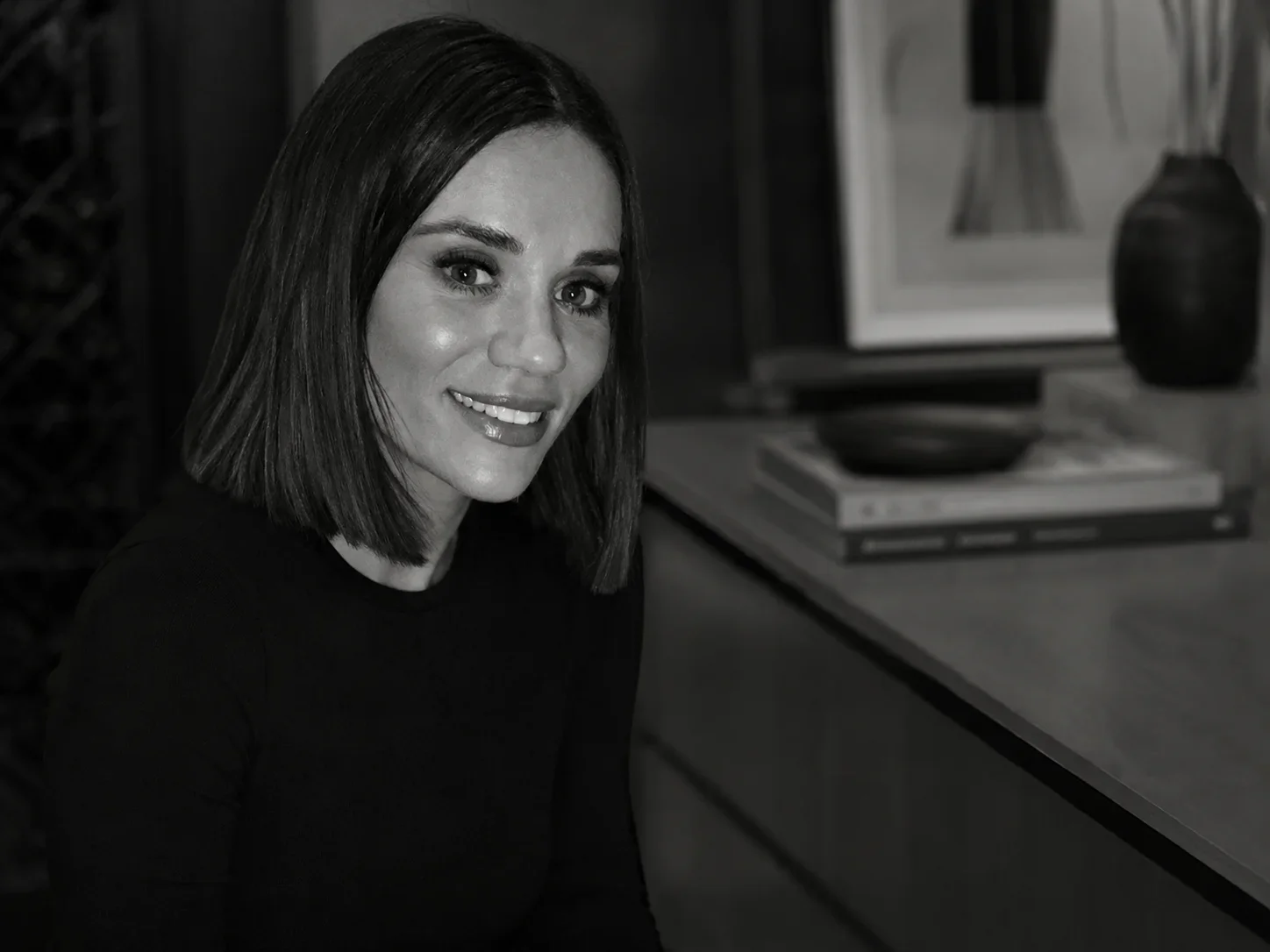 Black and white photo of a smiling woman with shoulder-length hair, sitting at a desk with a stack of books and decorative items in the background.