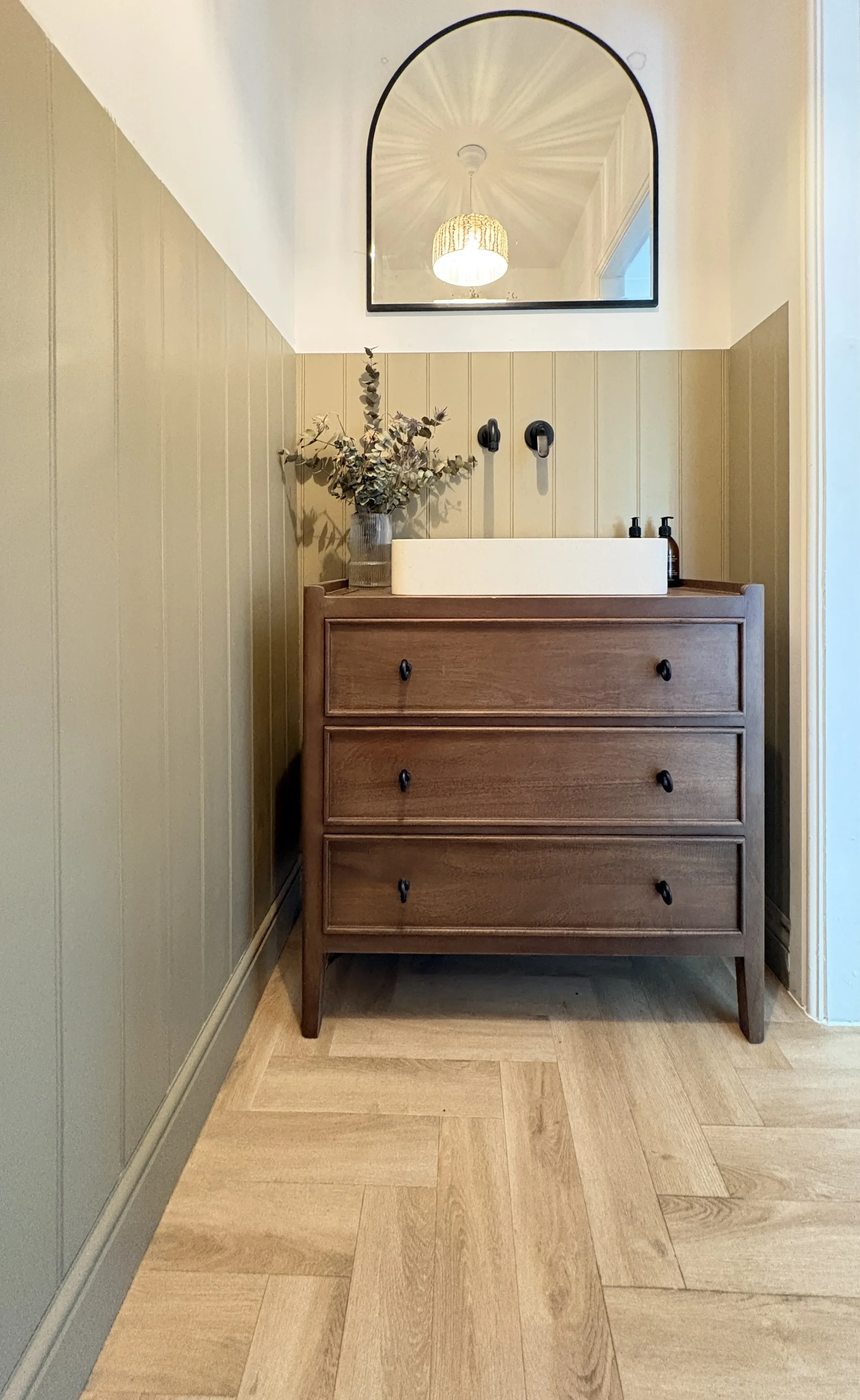 A wooden dresser with four drawers, topped by a white rectangular basin, a vase with dried eucalyptus, and two black bottles. Above, a mirror reflects a hanging light fixture in a room with white and beige walls and wooden flooring.
