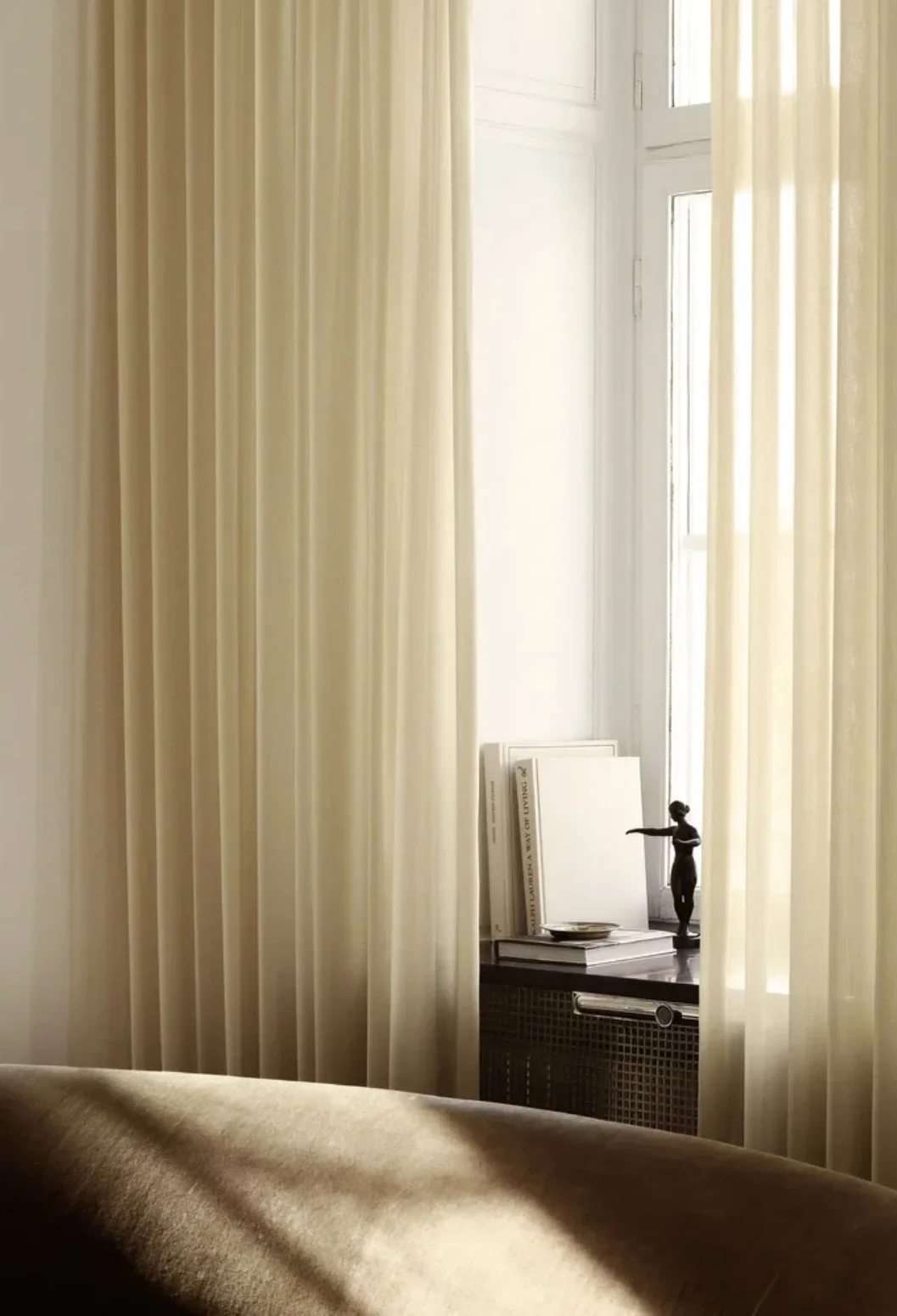 A corner of a room with beige curtains, a window letting in natural light, and a black sculpture of a woman on a ledge with books and decorative items.