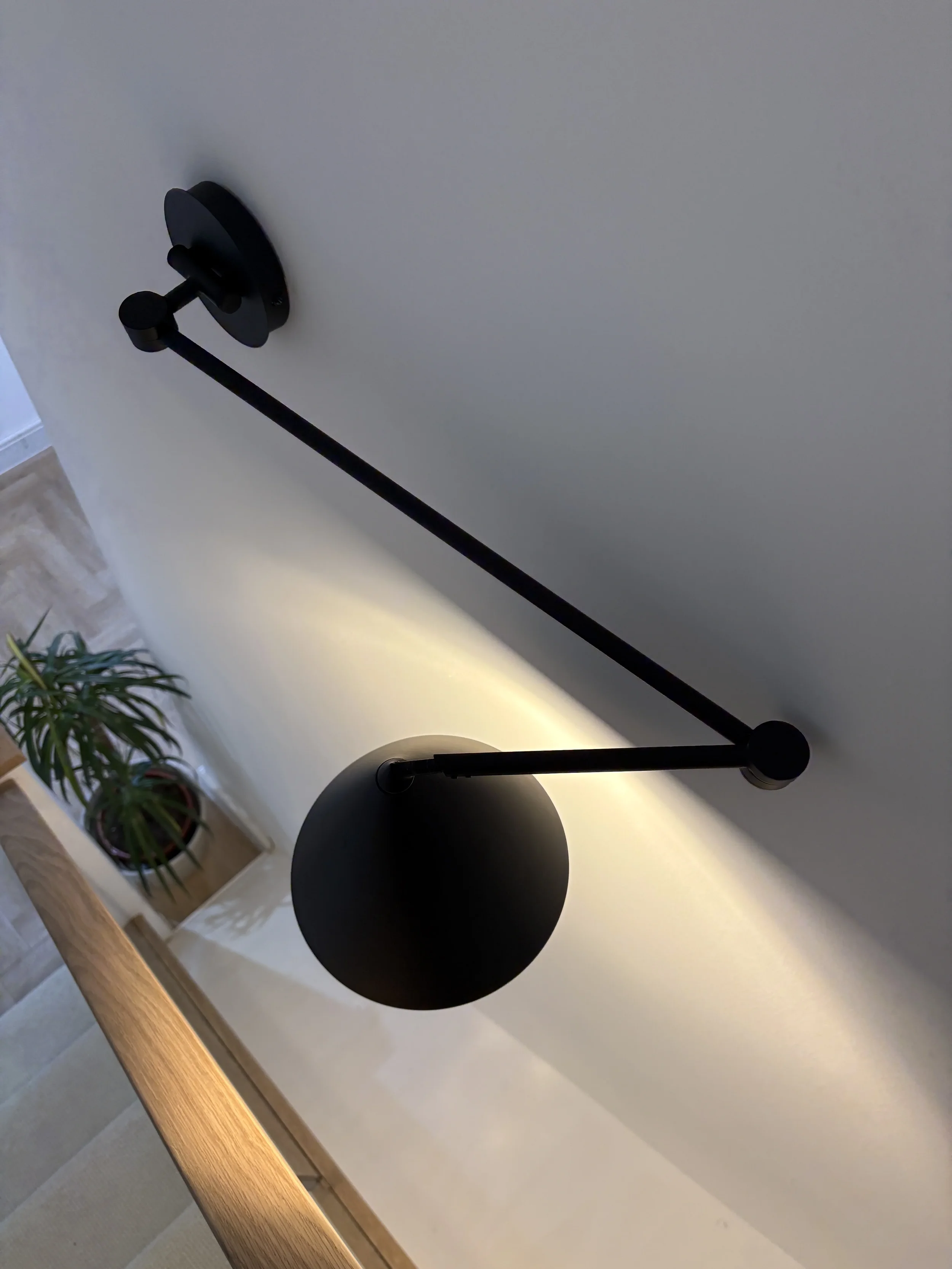 Close-up of a black adjustable desk lamp with a round shade, mounted on a wall above a wooden surface, with a potted plant on the side.