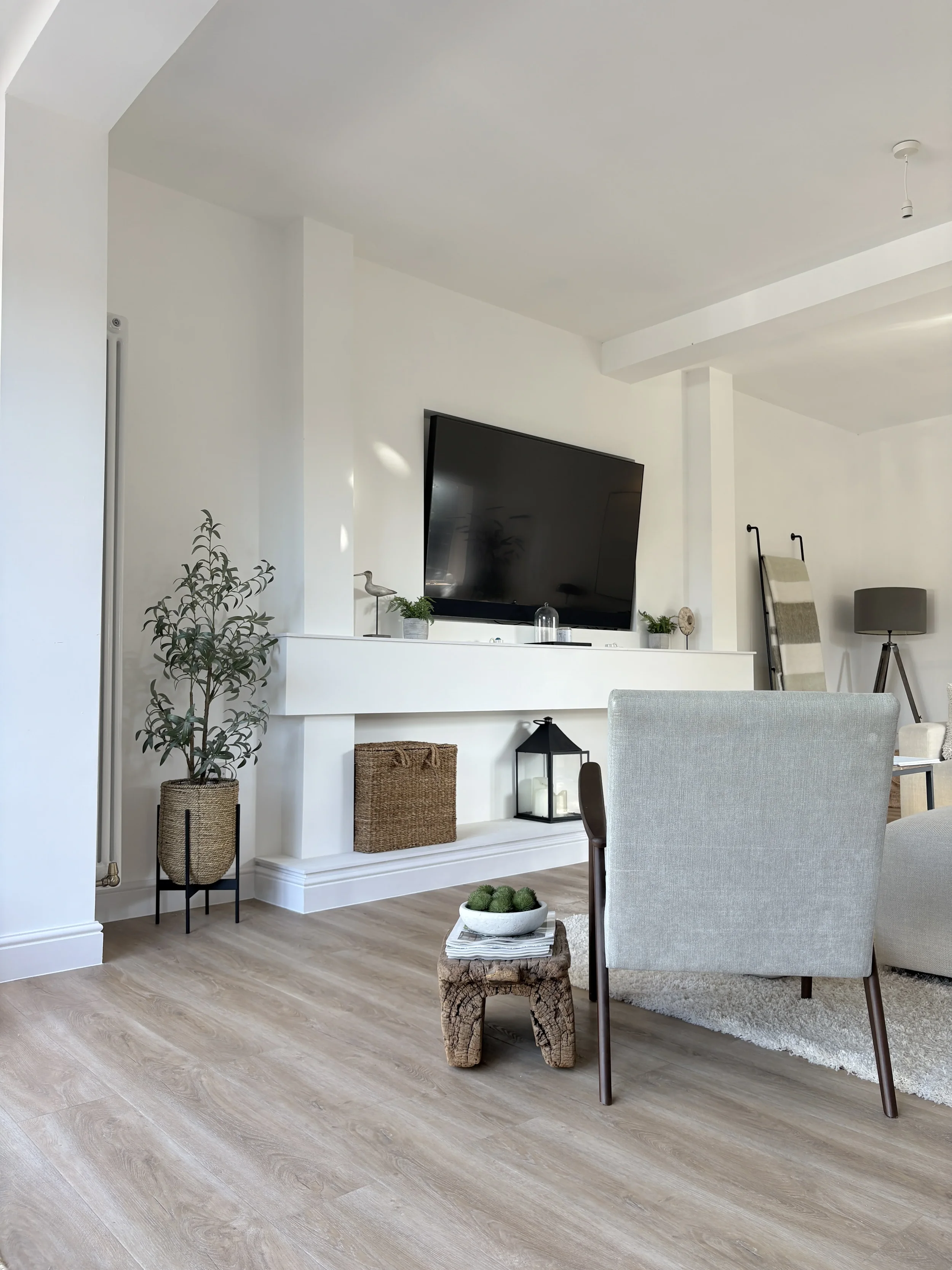 Living room with a mounted flat-screen TV, potted plants, a wicker basket, a lantern, a coffee table with a bowl of green decorative balls, a gray armchair, and a ladder leaning against the wall with a blanket, in a modern white and beige decor.