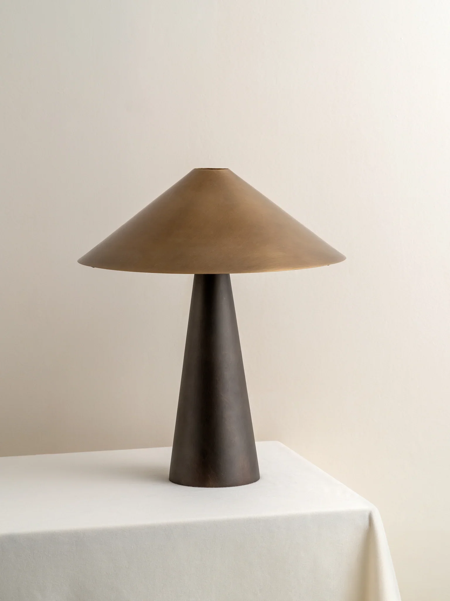 A dark wooden cone-shaped lamp with a beige lampshade on a white table against a plain white wall.