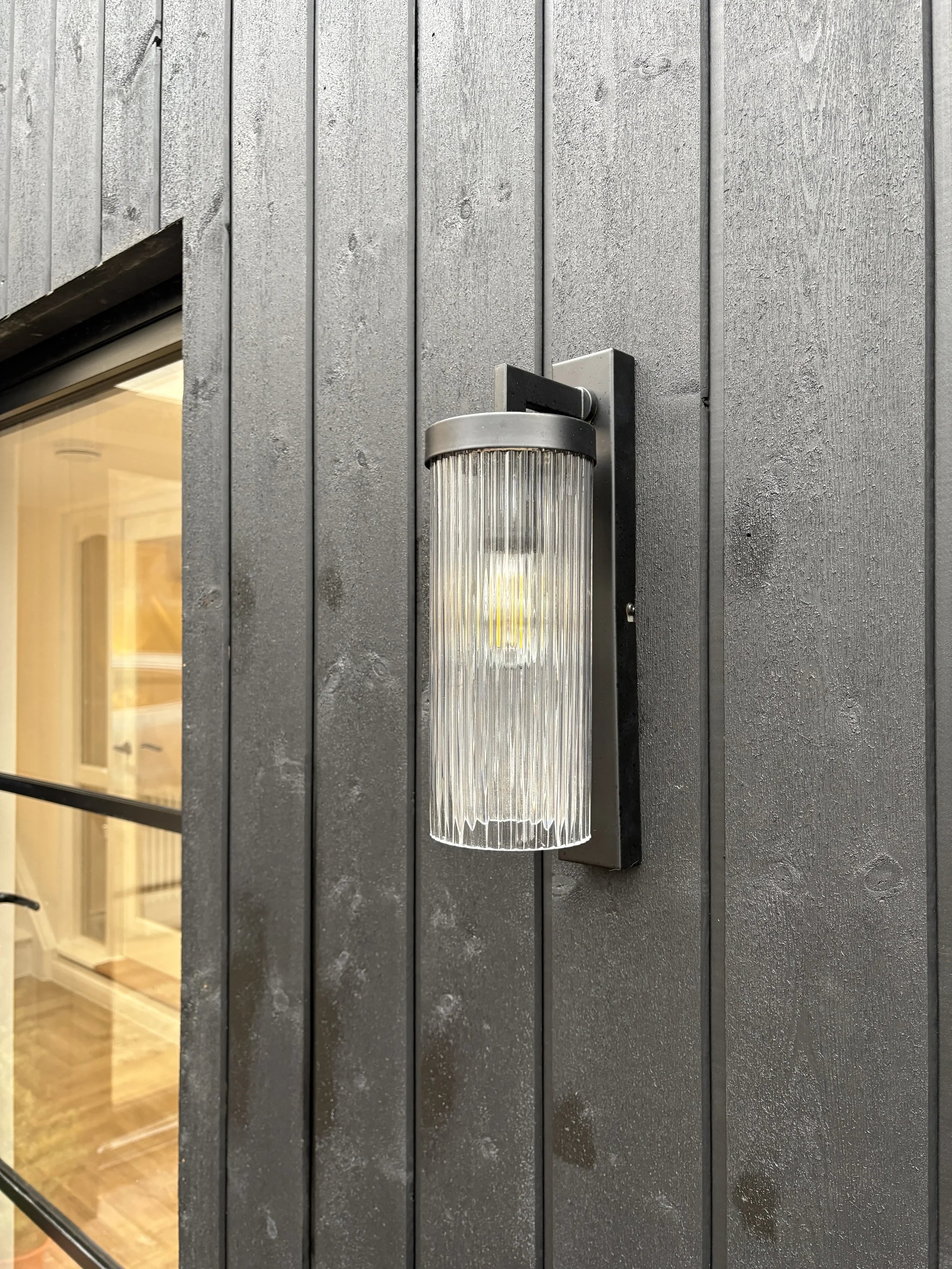 Contemporary exterior wall light mounted on black vertical timber cladding beside a steel-framed window, showcasing modern residential architecture and exterior detailing.