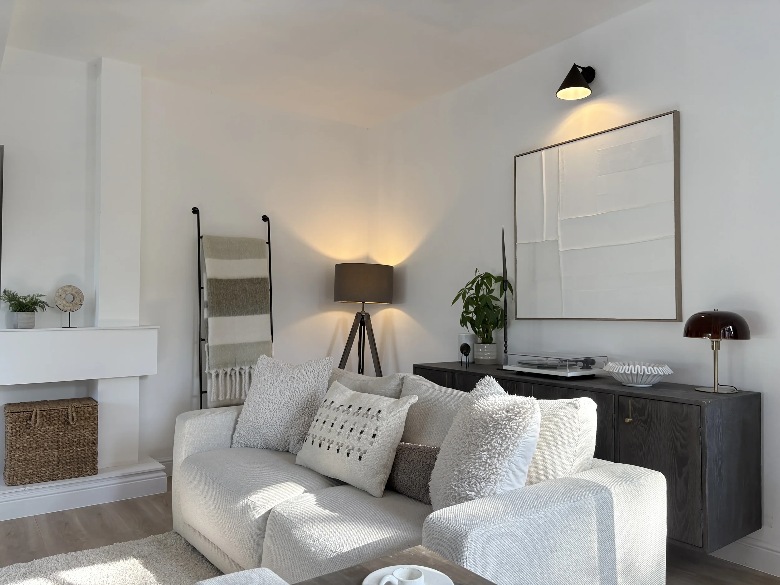 Living room with white sofa, decorative pillows, wall-mounted black lights, potted plant, wall art, wooden cabinet with turntable, and various decorative objects.