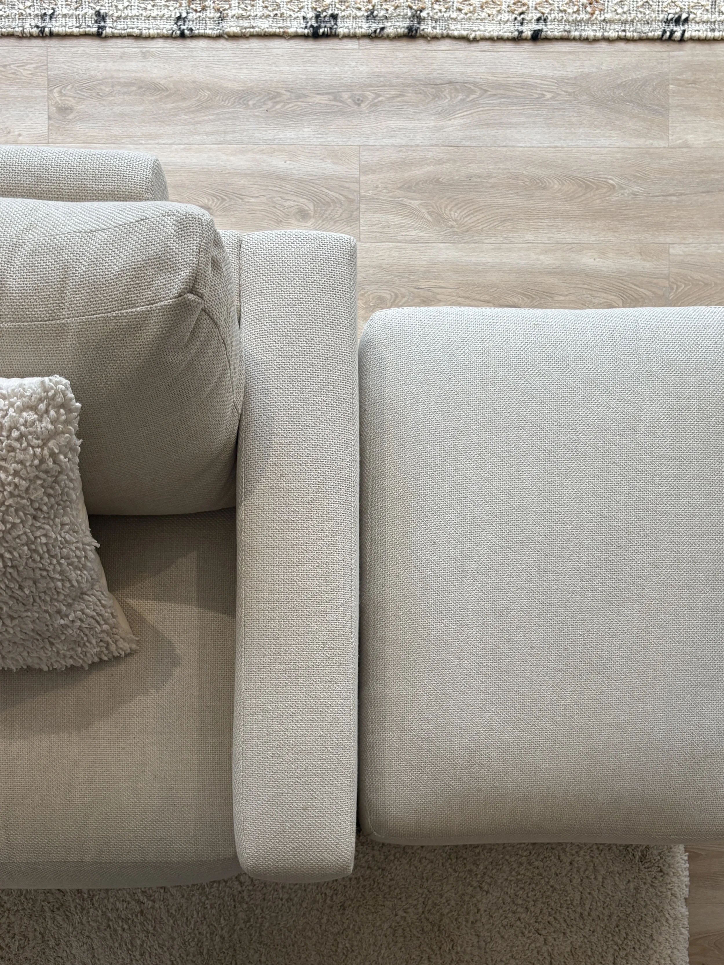 Top-down view of a beige sofa with a textured pillow and a blanket, positioned on a beige rug with a hardwood floor in the background.