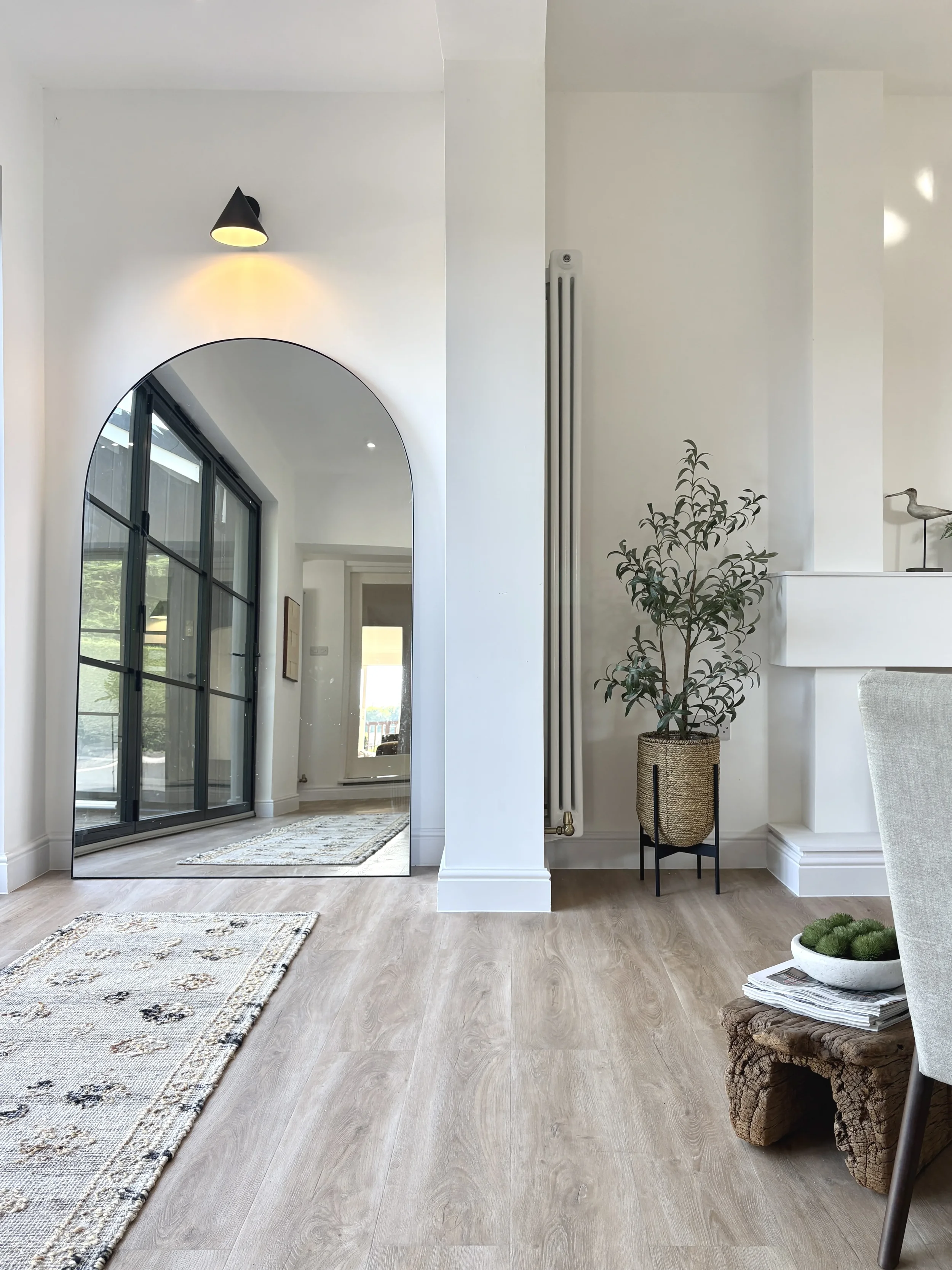 A modern living space featuring a large arched mirror reflecting a sliding glass door, a small potted plant, a rustic wooden side table with a bowl of plants, and a beige upholstered chair, with light-colored wood flooring and natural light streaming