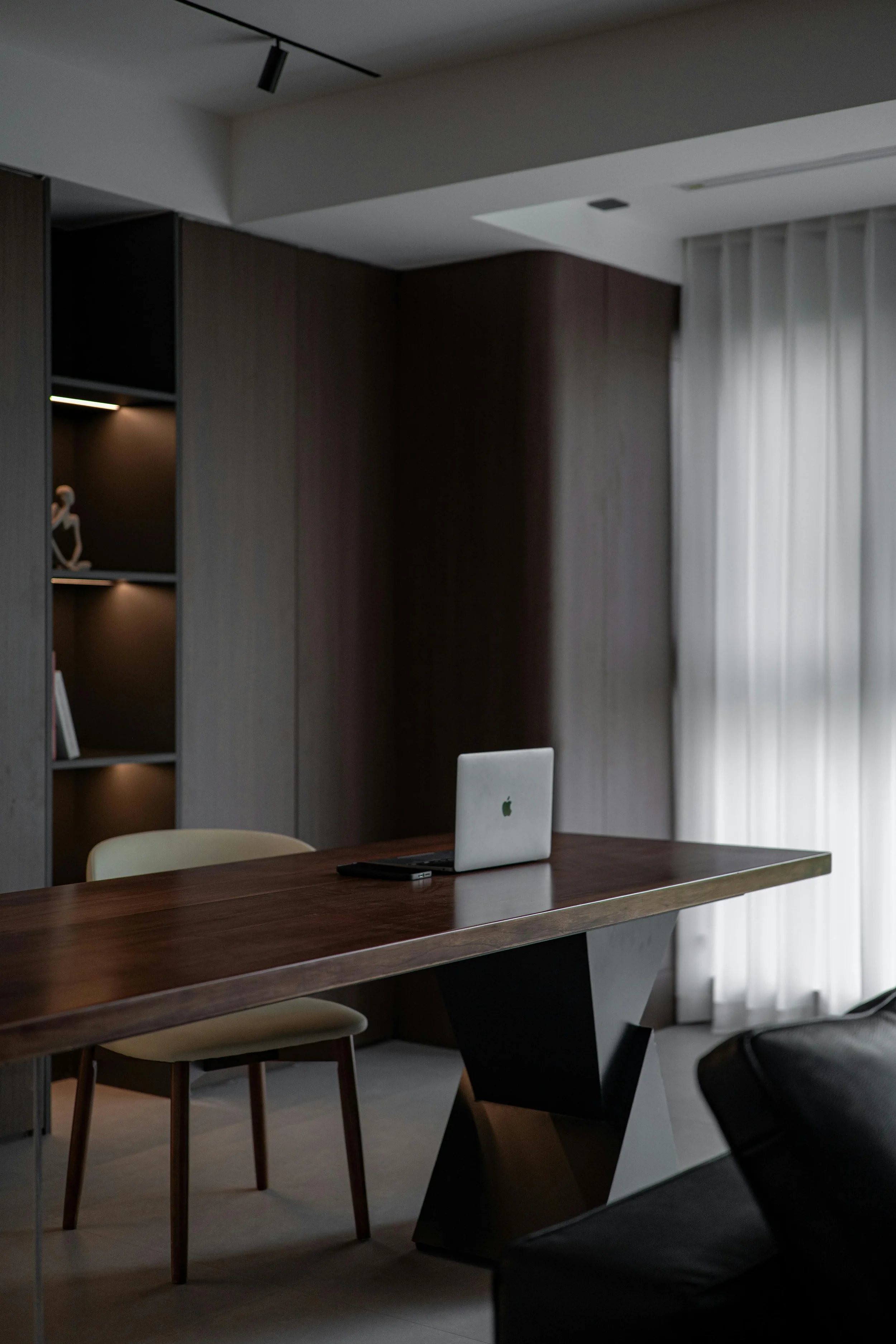Modern office space with a wooden meeting table, beige chairs, a silver MacBook laptop, and a dark leather chair. The room has dark wood paneling, light filtering through white curtains, and a ceiling light fixture.