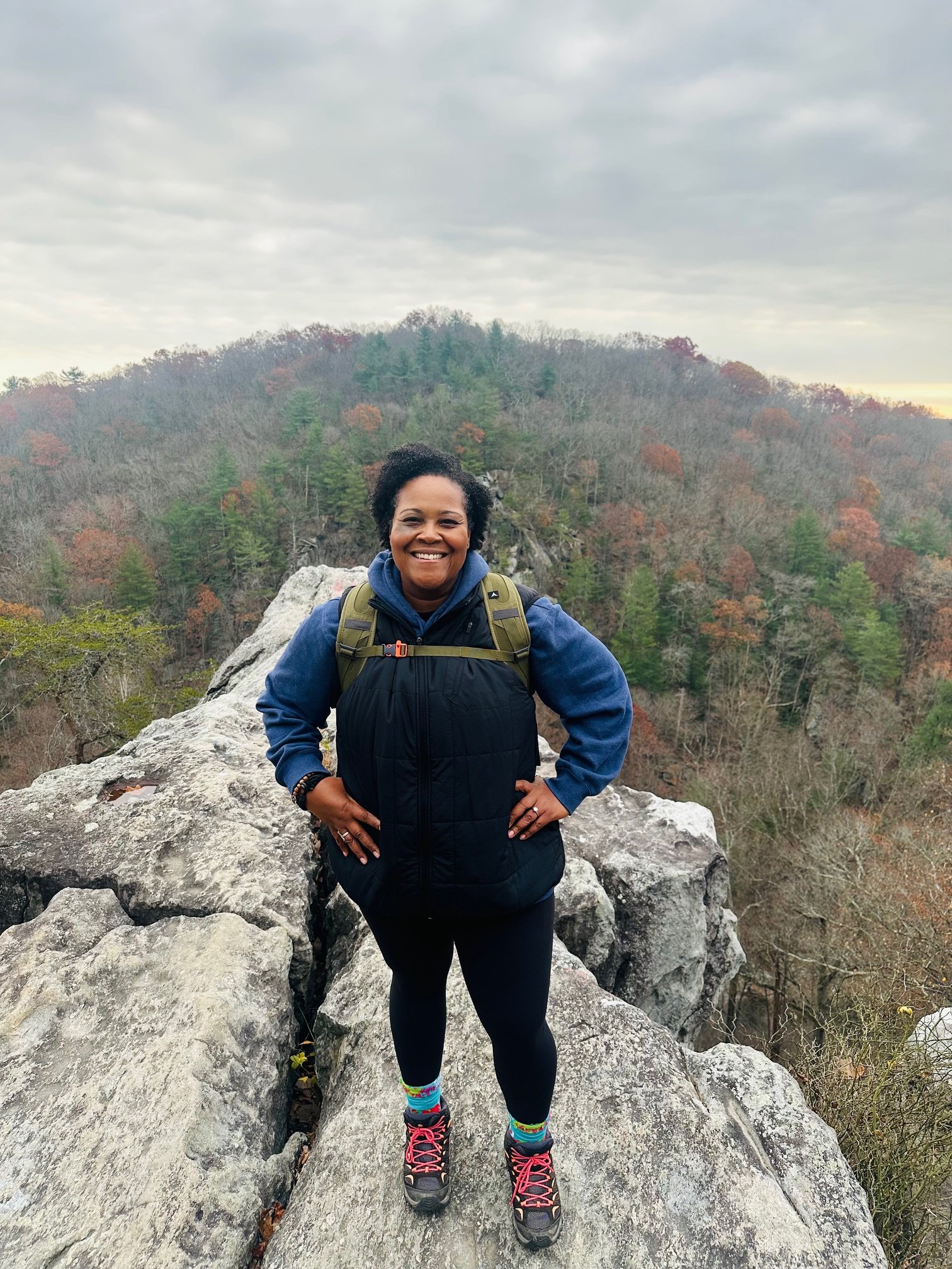 Nadji Kirby in outdoor hiking gear standing on a large rock with a forested mountain in the background during cloudy weather.