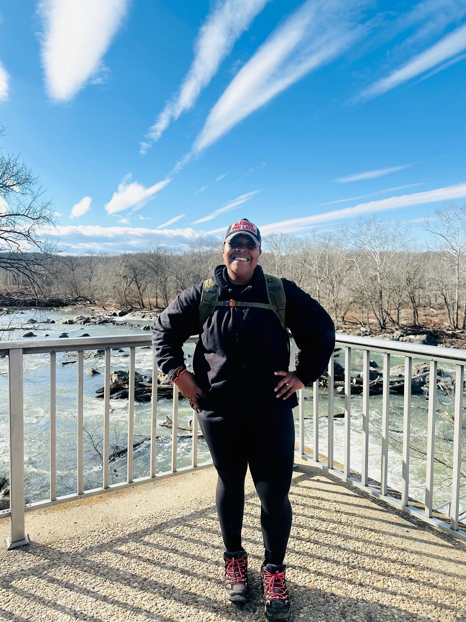 Nadji Kirby standing on a bridge by a river, smiling, wearing a black jacket, black leggings, hiking boots, and a cap, with leafless trees and a blue sky with clouds in the background.