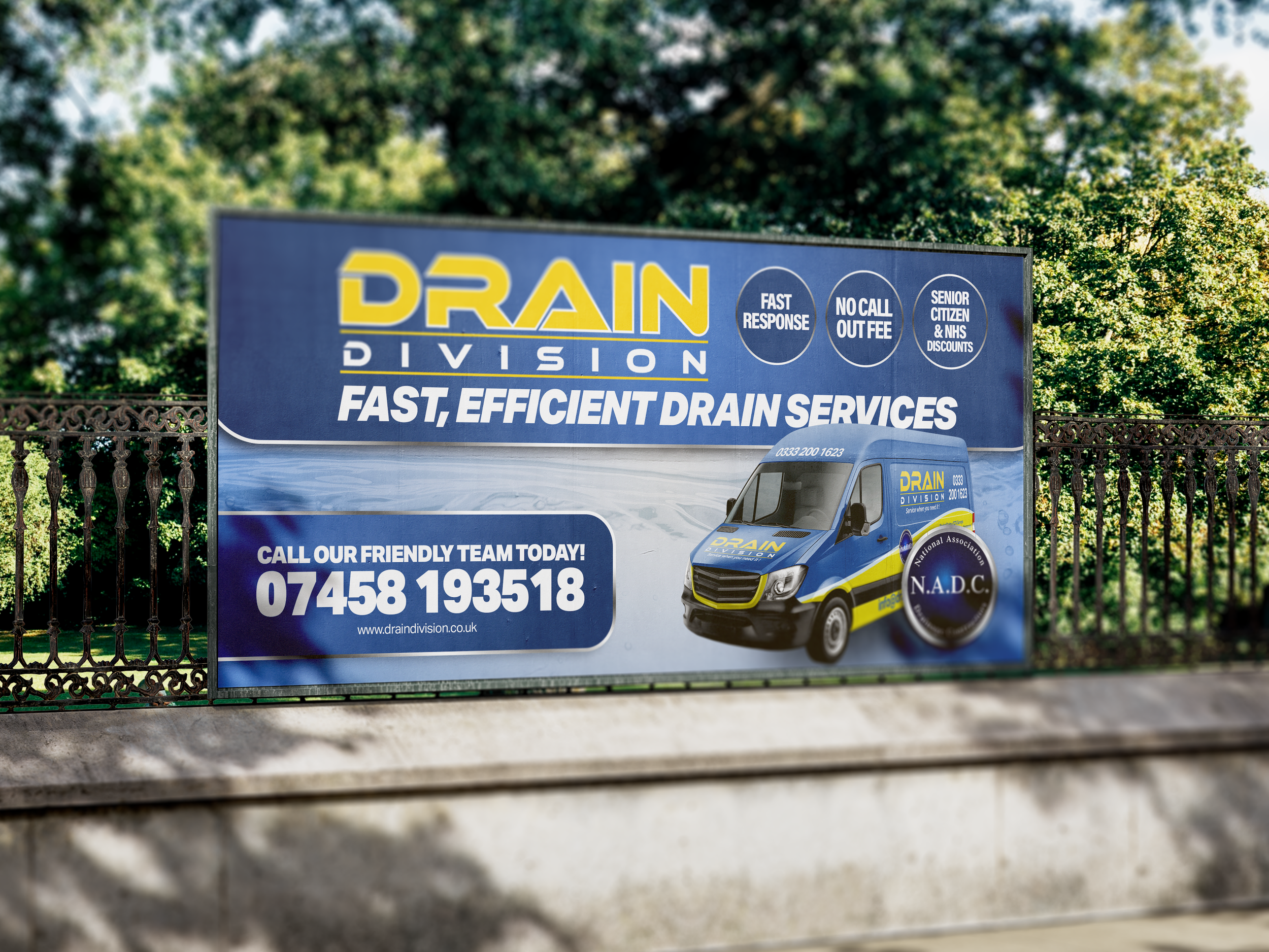 A large advertisement billboard for Drain Division services, featuring a blue and yellow van, contact information, and promotional tags, set against a backdrop of green trees.