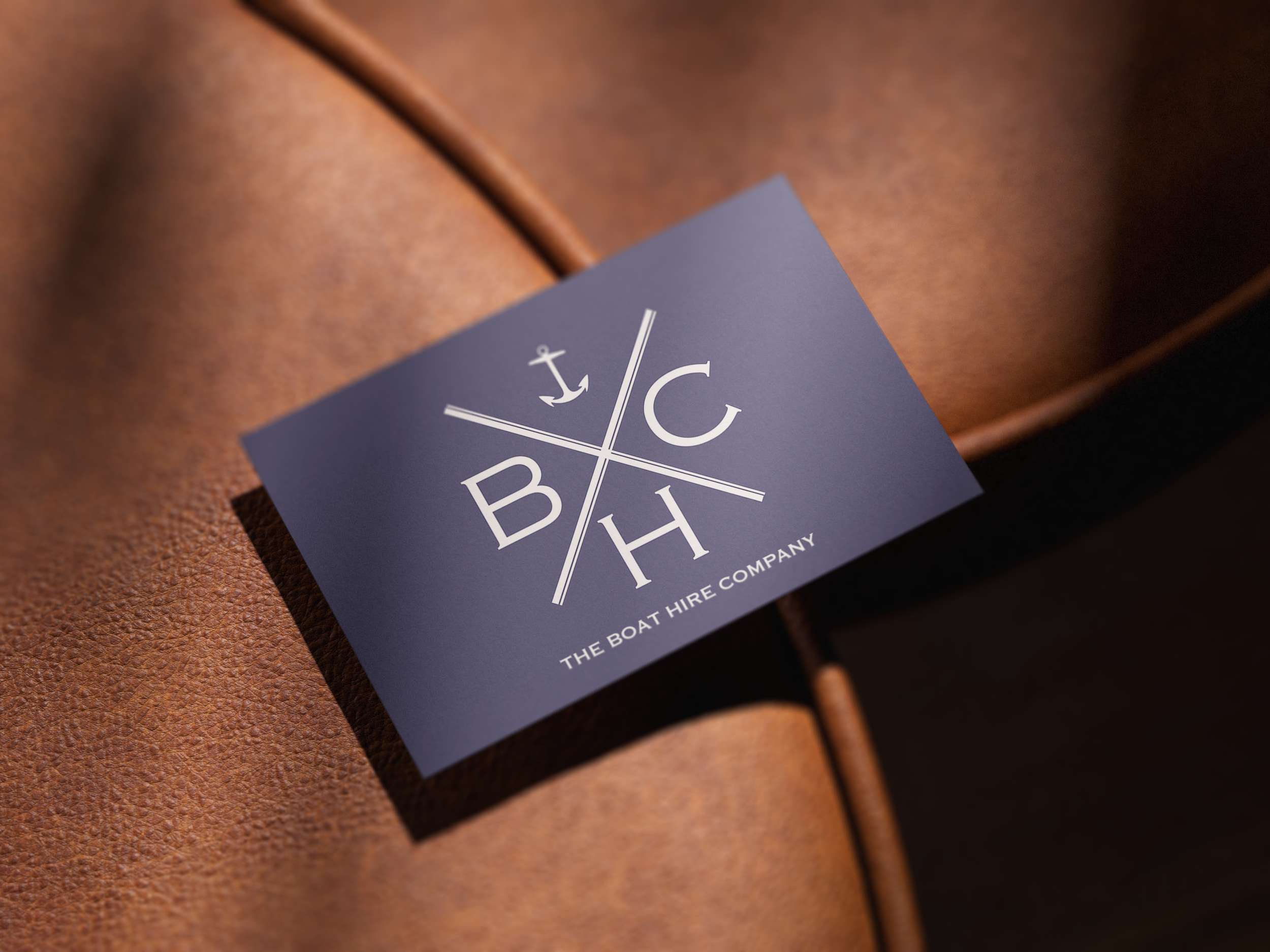 Close-up of a navy blue business card with white text and anchors in front of a brown leather surface, representing 'The Boat Hire Company'.