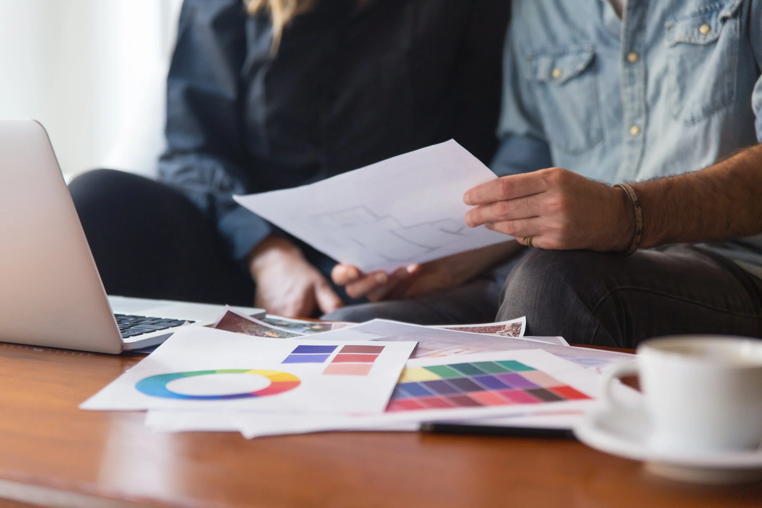 Two people sitting at a desk reviewing printed color and design samples, with a laptop and color charts on the table.