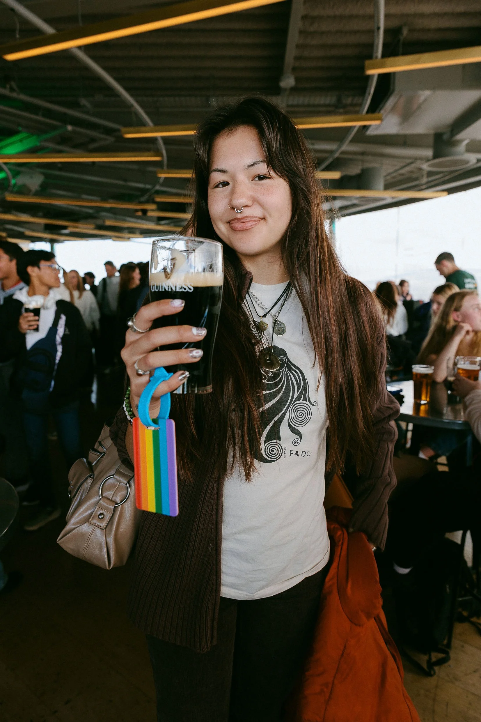 A young woman with long dark hair, wearing a white t-shirt with a spiral design and a brown jacket, holding a glass of dark beer with the Guinness logo, at a crowded indoor bar or restaurant.