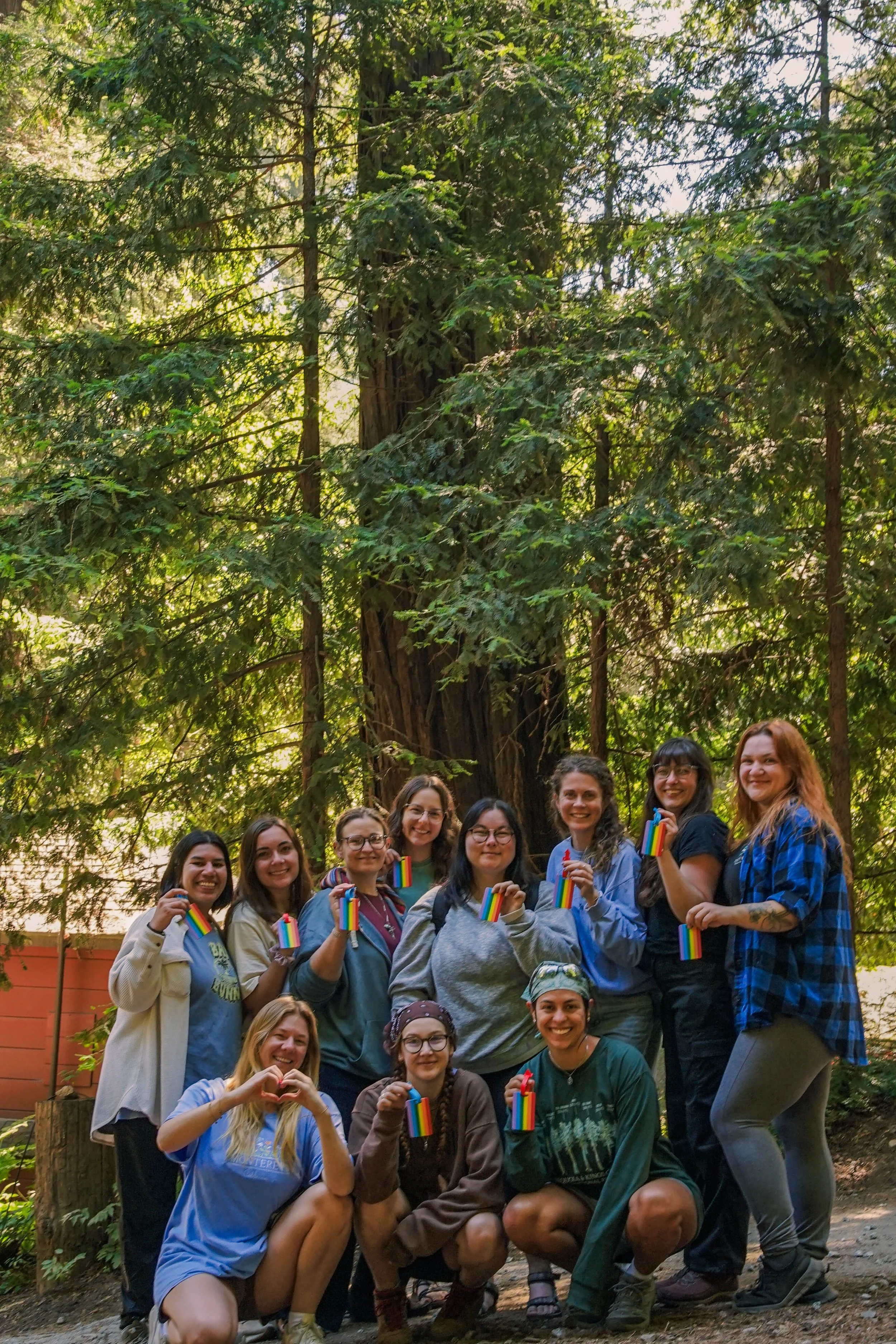 Group of smiling women holding rainbow pride flags in a forested outdoor setting.