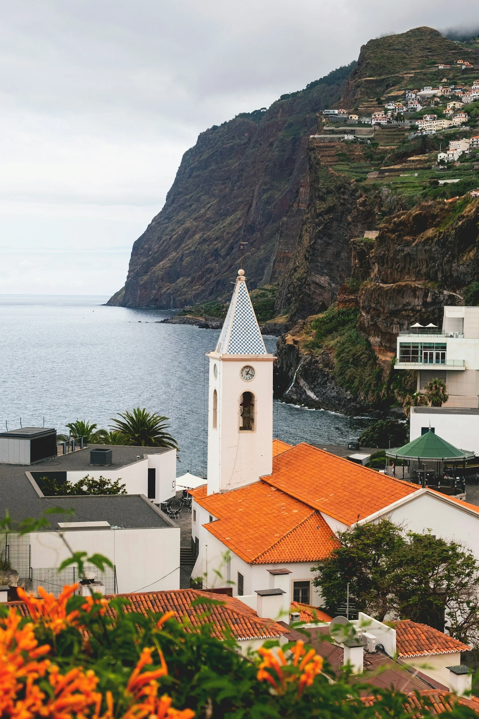 Coastal village built on steep hillside with houses, terraced gardens, and a prominent white clock tower with a blue and white checkered spire overlooking the ocean, beneath overcast sky, with vibrant orange flowers in foreground.