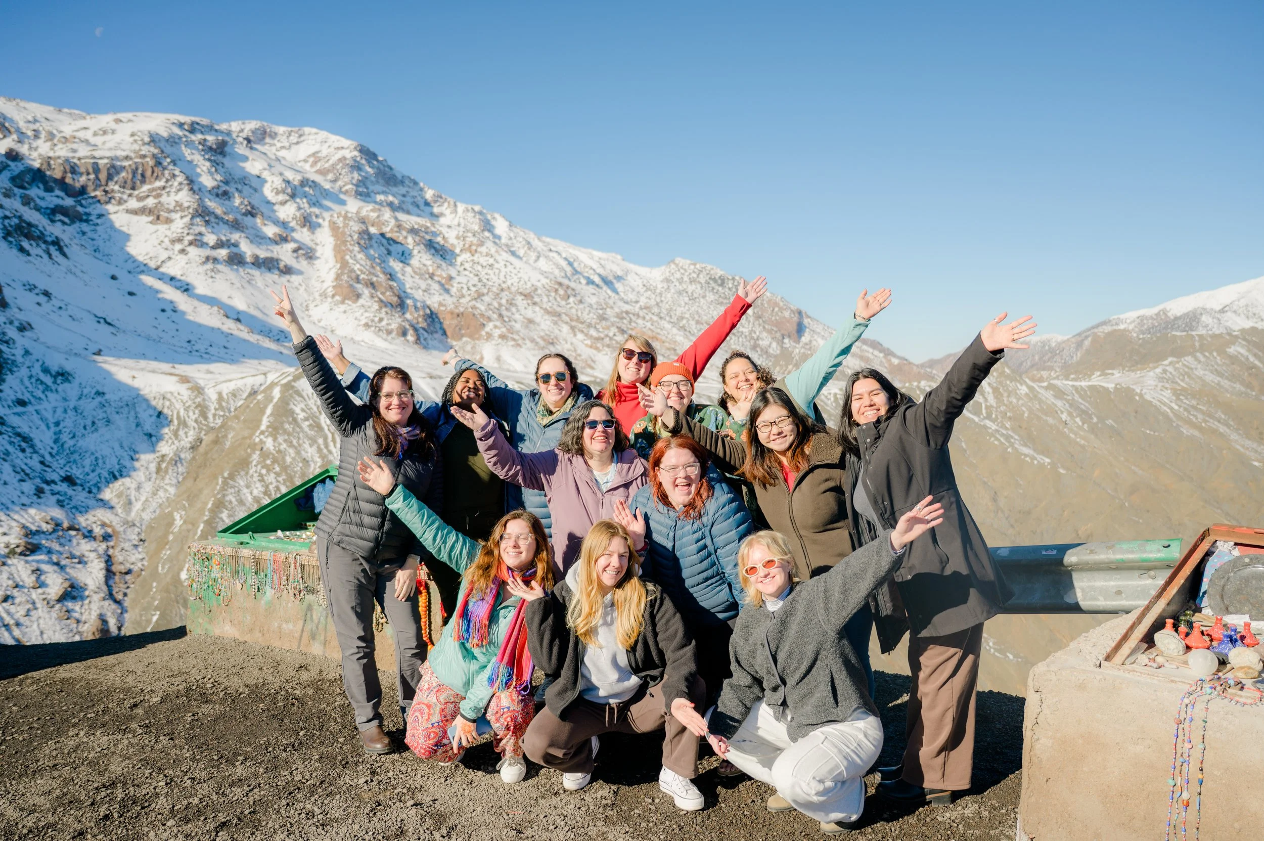 Group of cheerful people posing with their arms raised in front of a snowy mountain landscape under a clear blue sky.