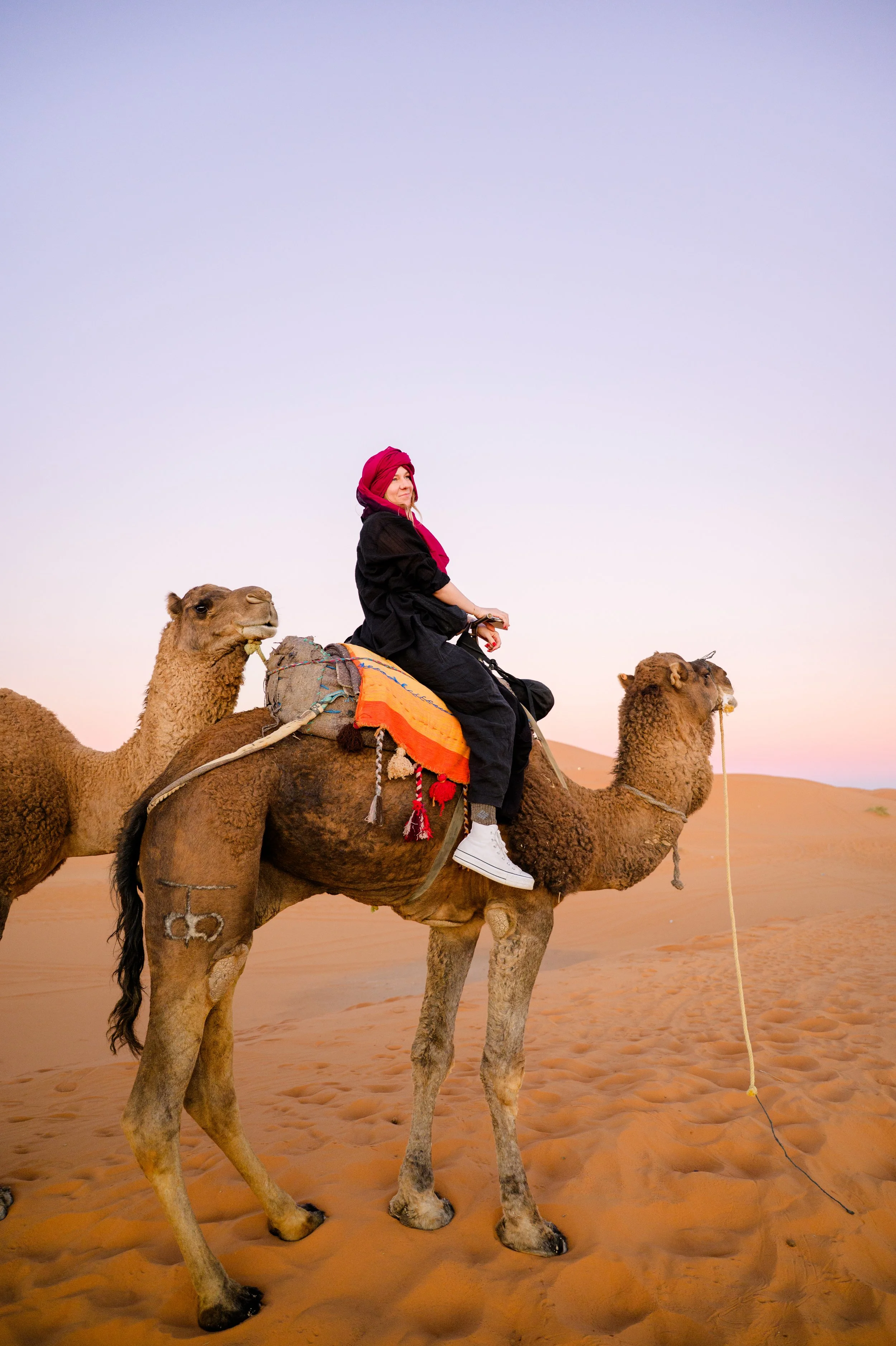 A woman in a black outfit and red headscarf riding a camel in a desert during sunset, with sand dunes in the background.