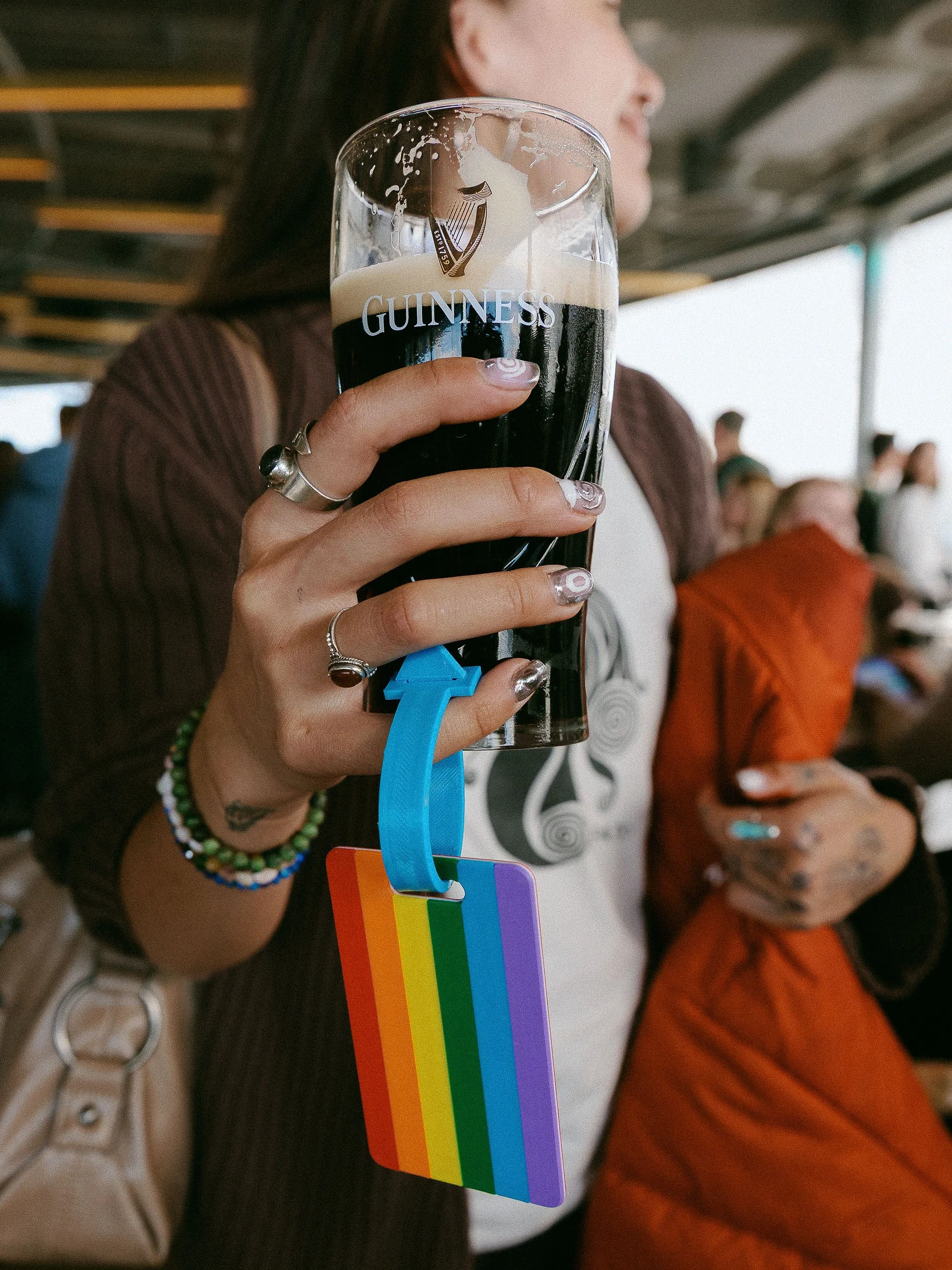 Close-up of a person holding a pint of Guinness beer with a rainbow pride flag pin hanging from their fingers. The background has people sitting at a table in a social setting.