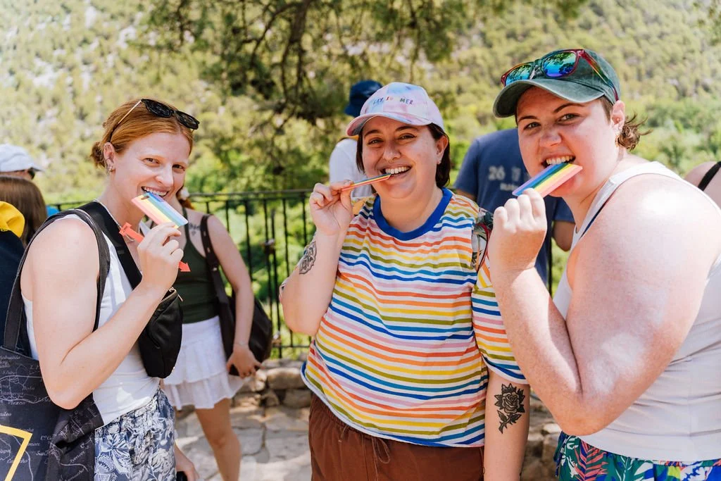 Three women smiling and eating rainbow-colored popsicles outdoors.