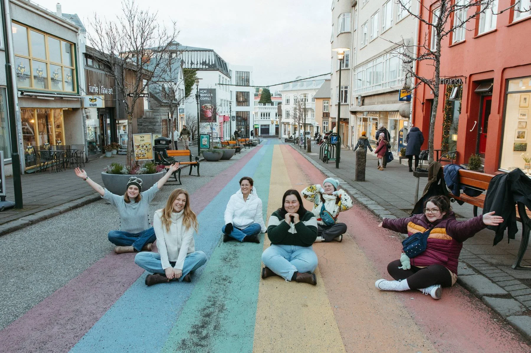 Group of people sitting on a colorful rainbow crosswalk on a city street, smiling and posing for the photo, with shops and pedestrians in the background.