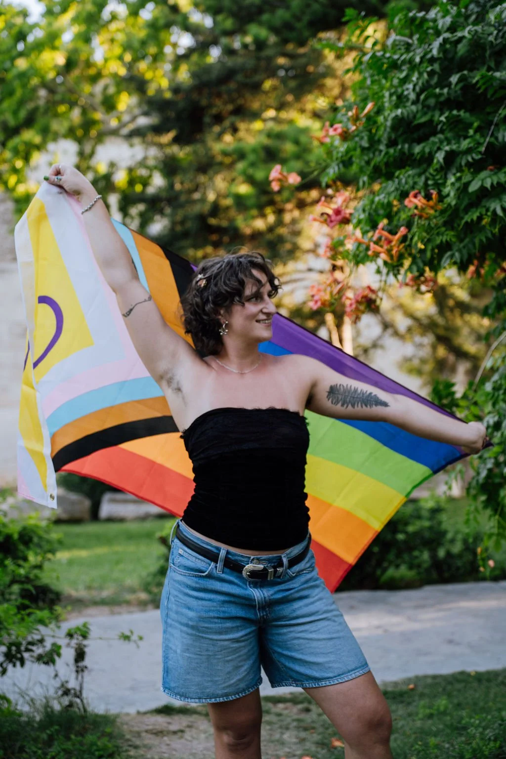 Woman standing outdoors in a park holding a rainbow pride flag, wearing a black strapless top and denim shorts, with trees and flowering bushes in the background.