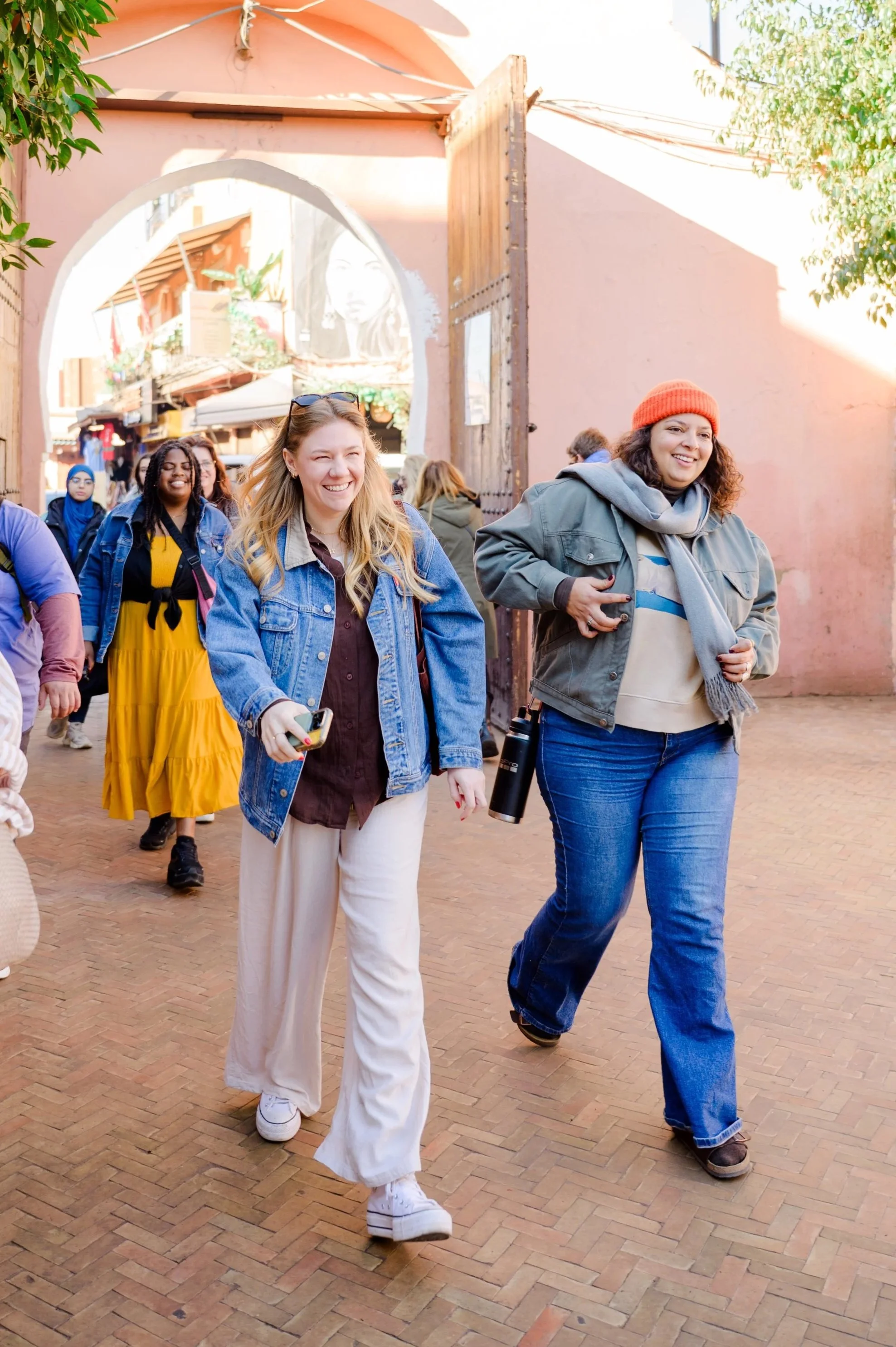 A group of diverse women smiling and walking outdoors in a lively, colorful area with pink walls and a mural of a woman’s face in the background. They are dressed casually, some holding phones or water bottles.